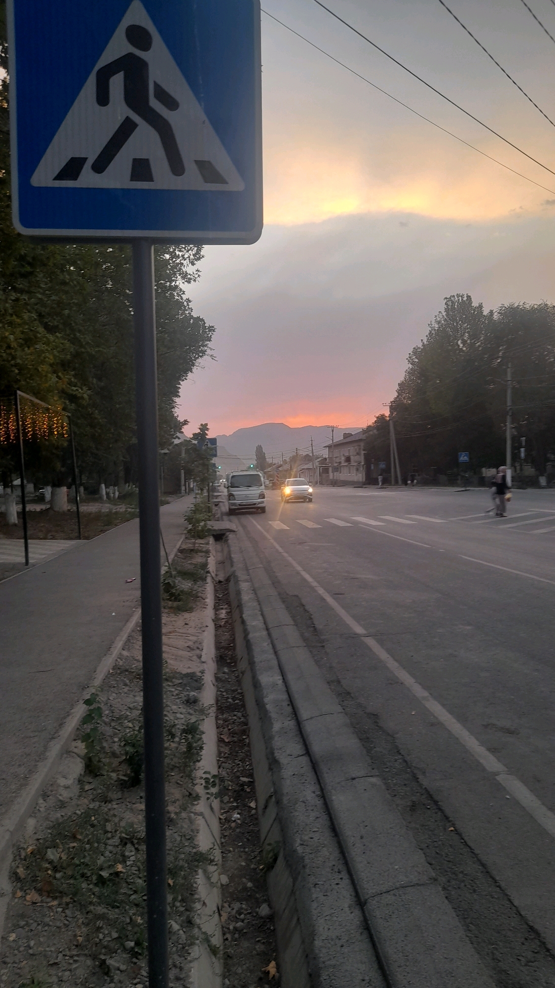The image depicts a street scene in Osh City, Kyrgyzstan, during sunset. A pedestrian crossing sign is prominently displayed on the left side of the frame. The sky is illuminated with warm orange and yellow hues from the setting sun, casting a soft light over the landscape. Trees line the sidewalk on the left, some adorned with decorative lights. A road runs through the center of the image, with cars and a motorcycle visible. In the distance, mountains form a backdrop against the colorful sky. The overall atmosphere is serene and peaceful, capturing a typical evening in the city.