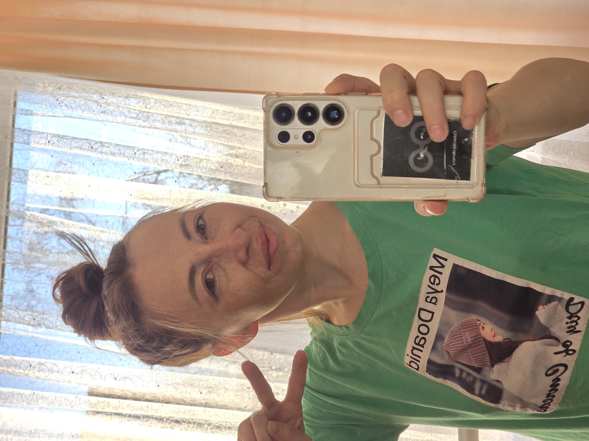 This selfie, taken in Chaikovsky, Russia, depicts a woman holding a smartphone and making a peace sign with her left hand. The image is captured in a mirror or reflective surface, evident from the reversed text on her t-shirt. The woman has a light complexion with visible freckles and her hair is pulled back into a bun. She gazes towards the camera with a slight smile, conveying a casual and positive mood.
She is wearing a green t-shirt featuring a printed image of a person, likely a woman, wearing a knitted beanie hat and a light-colored sweater. Vertical lines of reversed text are visible on the t-shirt; one line appears to read "ИМЭЛЯ ЭОСЫИЯ" and the other "Deal of Gmensis," suggesting a mix of Cyrillic and Latin alphabets or a highly stylized font. The smartphone, held in her right hand, is light-colored with a clear protective case, showcasing its distinctive multiple camera lenses (three large and two smaller ones), characteristic of a high-end model like the Samsung Galaxy S Ultra series. A black card or object is inserted into a slot on the back of the phone case, with some faint, unreadable white text.
The background reveals an indoor setting with abundant natural light streaming through a window. Above the window, an orange-toned roller blind is visible. The window itself is covered by a sheer white curtain adorned with a decorative patterned design. Beyond the window, a clear blue sky suggests sunny weather, indicating the photo was taken during the day, likely morning or afternoon.