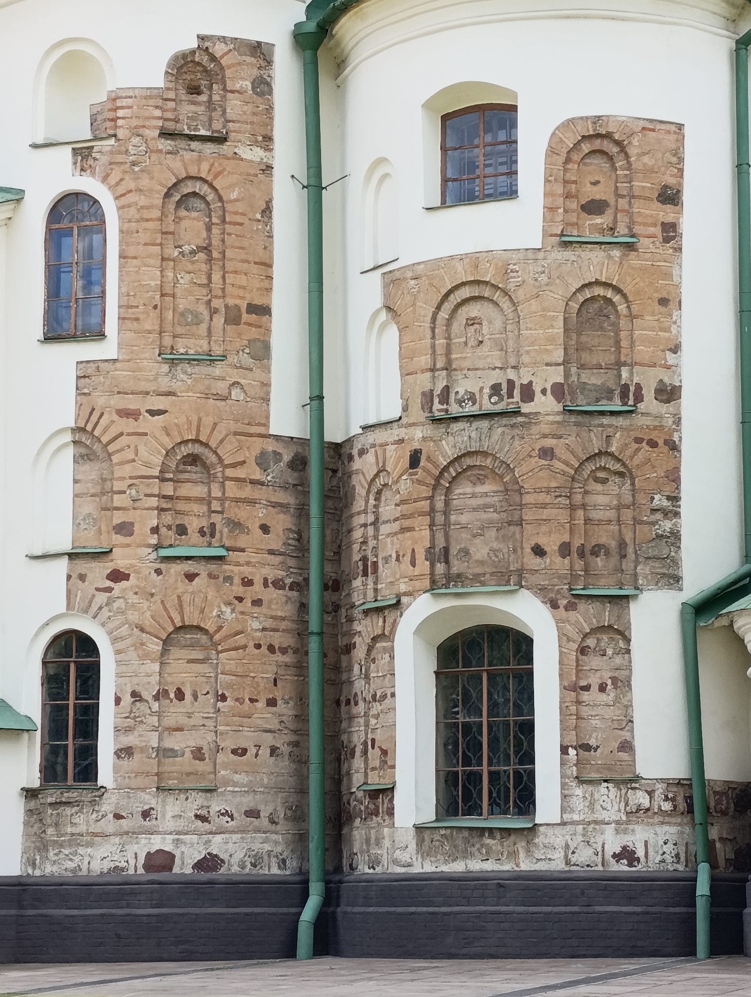 The image shows a close-up view of the exterior wall of a building in Kyiv, Ukraine. The wall is constructed with a combination of white plaster and weathered, reddish-brown brickwork. The brickwork features decorative arched niches, some of which are empty, while others appear to have been filled or altered over time. Several windows are visible, some with arched tops and metal grilles, and others with simpler rectangular frames. A prominent green downspout runs vertically along the white section of the wall. The overall impression is of an old, possibly historical building with varied architectural elements. The specific location in Kyiv is not identifiable from the visual information presented.