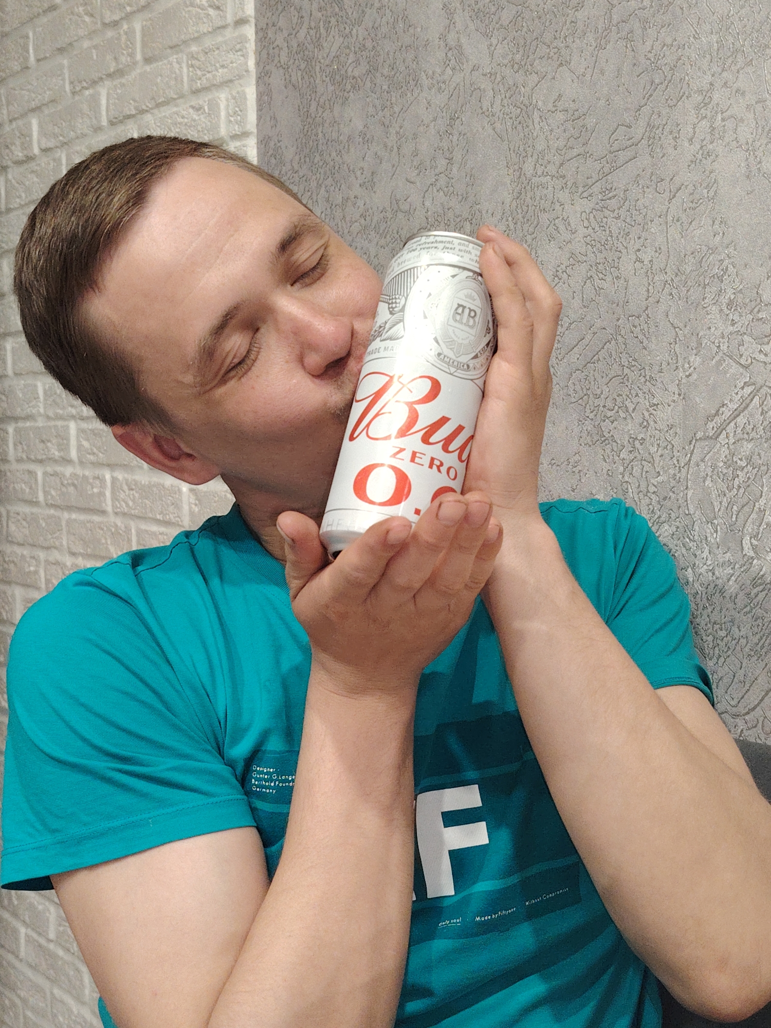 In Chaikovsky, Russia, an indoor shot captures a man holding a white can affectionately against his face. His eyes are closed, and he displays a soft, content expression. The man has short brown hair and is wearing a teal or turquoise t-shirt.
He holds the can with both hands, with the prominent red lettering "Bud ZERO" clearly visible on its side. Other text on the can includes a circular "AB" logo, "AMERICA," and "TRADE MARK," along with fragmented words like "Ahment, and s," "reare, just with," and "MADE MA."
The background consists of a light-colored, possibly painted, brick-patterned wall to the man's left, and a textured grey wall to his right. Visible text on the man's t-shirt includes "Designer : Gunter G. Lange Berthold Foundr Germany," a large capital "F," and phrases such as "enly coul," "Made by Filtyone," and "Wilbaut Compromiss." The lighting suggests an interior setting, likely during the day.