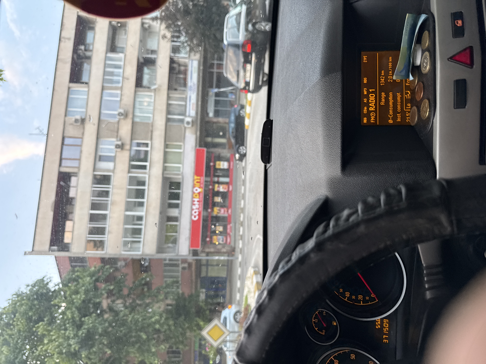 The image captures a street scene in Asenovgrad, Bulgaria, viewed from the interior of a vehicle. The specific location is the intersection of ulitsa Byala Reka and bul. Bulgaria, with the "CASHPOINT" store located at bul. Bulgaria 59 directly ahead.
Inside the vehicle, the lower portion of the steering wheel and the dashboard are visible. The instrument cluster on the left displays an odometer reading of 371509 km, a fuel gauge showing a low level, and a tachometer at zero RPM. The central digital display indicates "FM RADIO 1", a "Range" of 1342 km, an average consumption of "2.0 Lt./100 km", and an instantaneous consumption of "0". Below this display, a collection of Euro coins (including 10, 20, 50 cents, and 1 Euro denominations) and a folded Bulgarian Lev banknote are placed in a small black holder. A red hazard light button is positioned beneath the display area.
Through the windshield, a multi-story building occupies the central background. At its ground level, a "CASHPOINT" store with a prominent red sign and yellow-illuminated interior is the focal point. To the left of the "CASHPOINT" store, the ground floor shows additional commercial spaces. Further left, a large, mature green tree partially obscures another building. In the immediate street foreground, a yellow diamond-shaped "priority road" traffic sign stands at the edge of a small, raised traffic island marked with yellow and white stripes on the curb.
Several vehicles are present on the street. A silver car, possibly a BMW, is observed turning left behind the priority sign. A dark sedan, potentially an Audi A4, is visible further down the street past the "CASHPOINT" store. On the right side of the frame, a dark grey Audi A6 C6 Avant (station wagon) is parked or moving, displaying a Bulgarian license plate. The sky above is partially cloudy with patches of blue.