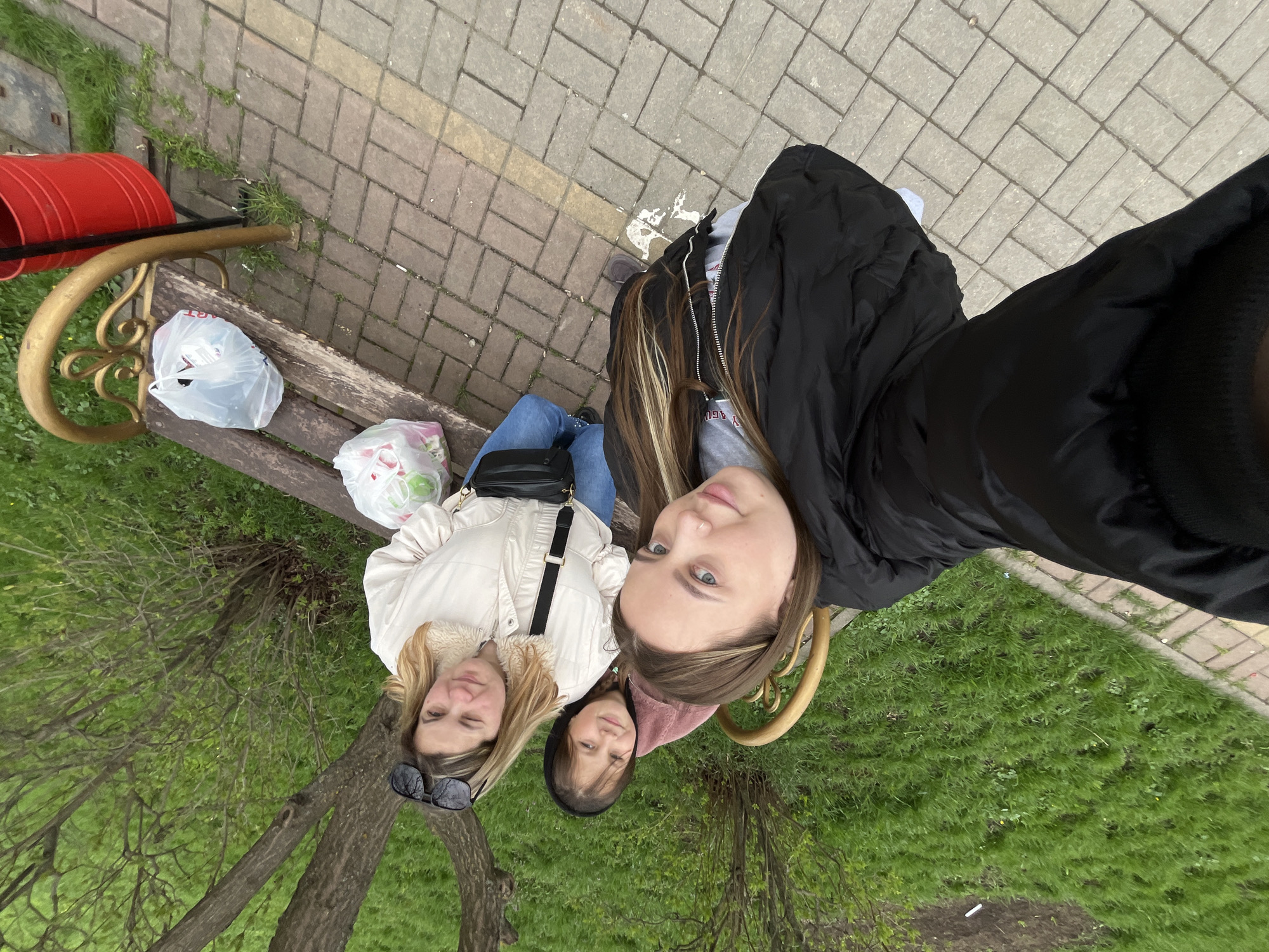 Three females are sitting on a park bench. One person wears a black jacket, another a light-colored jacket, and the third a dark-colored hooded sweatshirt. Plastic bags are on the bench next to them. The bench is next to a brick pathway. A red trash can is to the left side of the bench. The background shows grass and trees. The photo was taken in Novopokrovskaya, Russia.