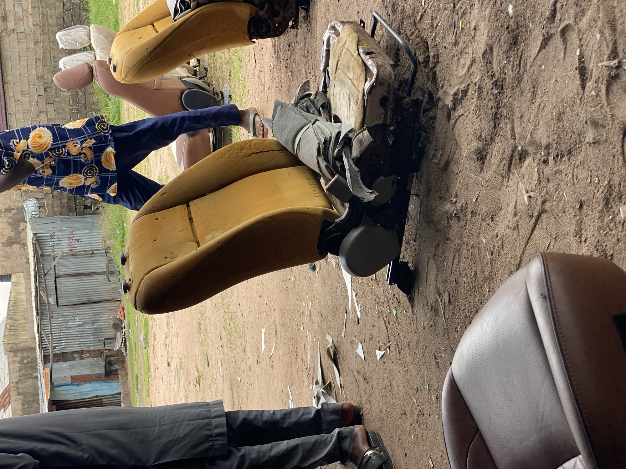 The image depicts an outdoor scene in Jalingo, Nigeria, likely a workshop or junkyard for automotive parts. In the foreground, a brown leather car seat is visible, showing a perforated texture. Behind it, several car seats are scattered on the sandy ground. One seat, in particular, is a prominent yellow, with its foam exposed and damaged. It's mounted on a metal frame with visible mechanical parts. To the left, a person wearing a gray robe and black sandals is partially visible, standing beside a pile of debris. Further back, another individual is seen wearing a blue shirt with a vibrant, patterned design and dark blue pants, walking among other car seats, some of which are covered in white plastic. The background reveals corrugated metal structures, possibly makeshift buildings, and a rough brick wall. The ground is primarily sandy dirt with scattered debris, including pieces of paper and plastic. The overall impression is one of a functional but unkempt area dedicated to the repair or salvage of vehicle components.
