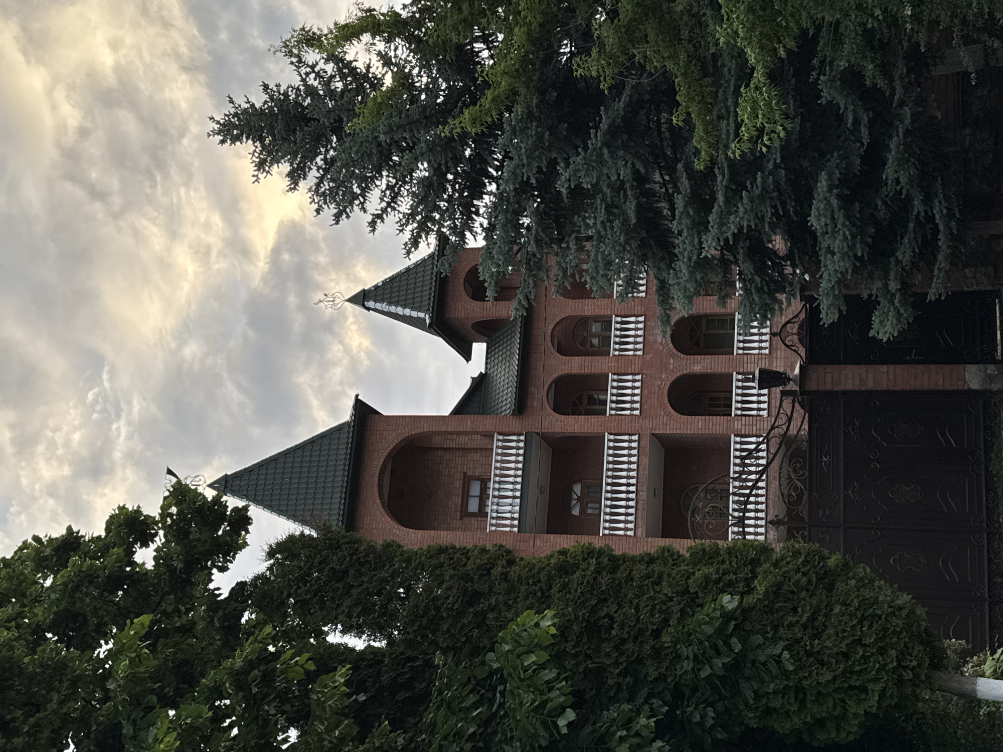 The image depicts a three-story brick building with arched windows and balconies, flanked by lush green trees. The building has two prominent towers with pointed, dark green roofs and decorative finials. A dark metal gate with ornate scrollwork is visible in the foreground, partially obscured by foliage. The sky above is cloudy with a hint of sunlight breaking through. The architectural style suggests a residential or possibly a small institutional building. The presence of mature trees and the building's design might indicate a location in a suburban or semi-rural setting. This building is located in Karachayevsk, Russia.