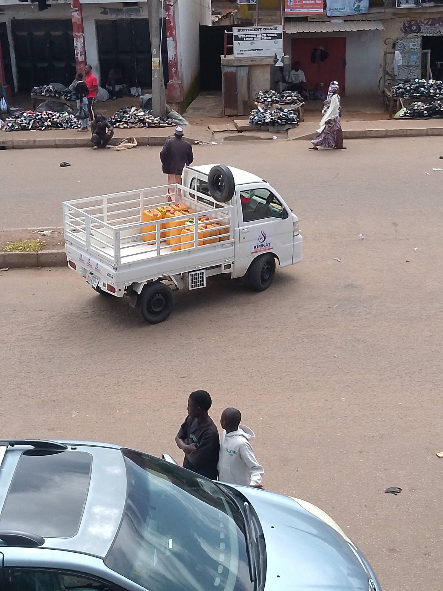 The image shows a street scene in Bukuru, Nigeria. In the foreground, a light blue car with a sunroof is parked. Two young men are standing near the car, looking to the right. Behind them, a white pickup truck is driving on the road. The truck's bed is filled with yellow canisters, possibly containing fuel or water. The truck has a tire mounted on its rear.
In the background, several people are visible near shops lining the street. One man in a red shirt stands by a table piled high with shoes. Another man in a dark jacket and cap walks away from the camera. A woman in a patterned dress and headscarf walks across the street. Piles of shoes are displayed on tables and the ground in front of the shops. Some buildings have signs, including one that reads "SUFFICIENT GRACE". The ground is paved with asphalt. The environment appears to be a busy marketplace.