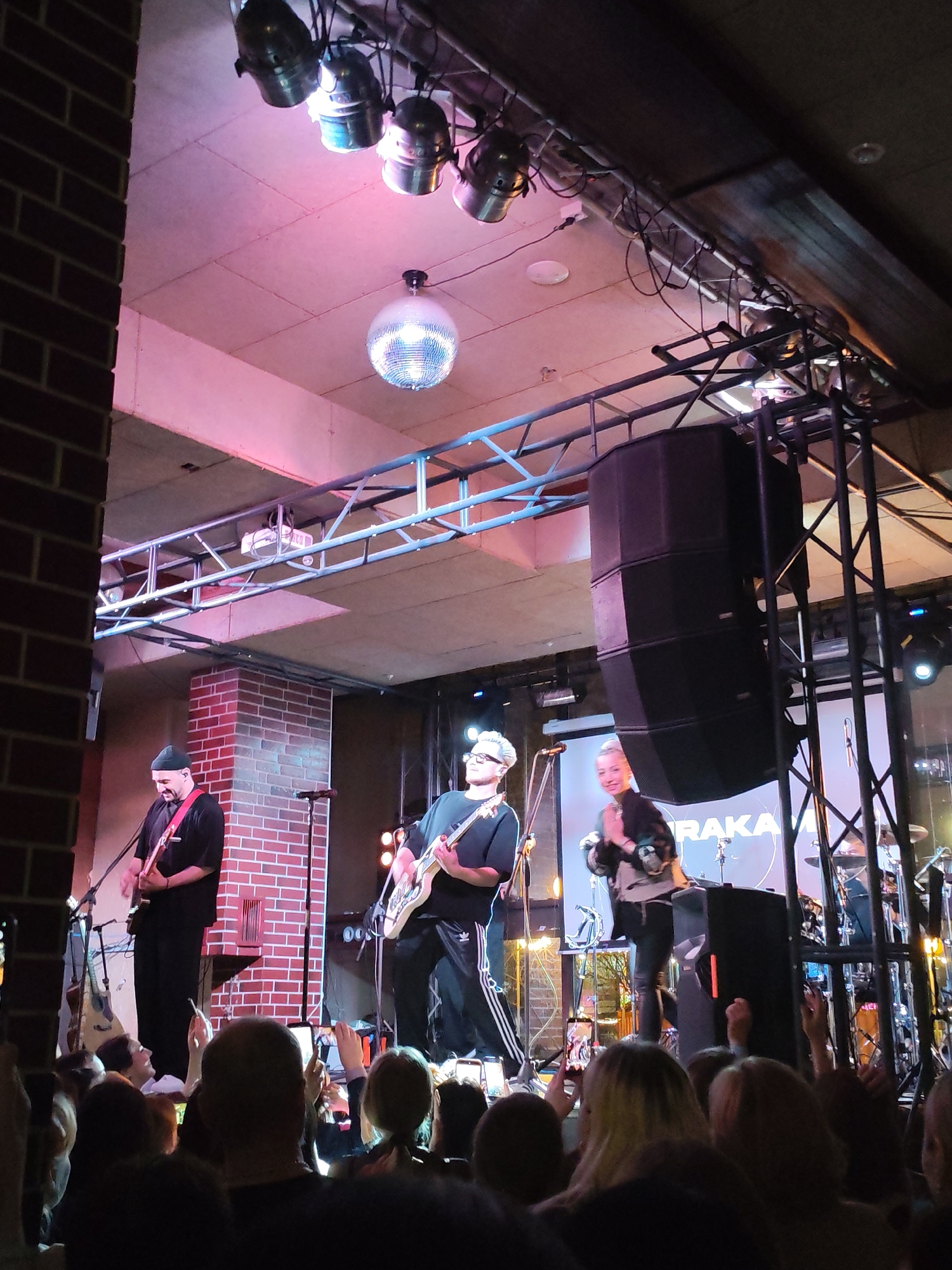 This image captures a vibrant indoor concert scene at what appears to be a club or music venue in Izhevsk, Russia. The perspective is from the audience, looking up towards the stage.
Three band members are performing on stage. On the left, a guitarist with a dark beanie and black clothing plays an electric guitar, standing near a brick pillar. In the center, another guitarist with glasses, wearing a black t-shirt and track pants, also plays an electric guitar. To the right, a third individual, likely the lead singer, with light hair, wears a dark patterned jacket and interacts with the audience. A drum set is partially visible behind them.
The stage is well-equipped with professional lighting and sound systems. A large stack of dark speakers is positioned on the right side of the stage. Overhead, metal trusses support various stage lights, including several spotlights and a prominent disco ball hanging from the ceiling, casting a festive glow. The ceiling itself is made of light-colored panels, illuminated with a soft pinkish light.
In the background of the stage, a large screen displays white text, with "RAKA" clearly visible, likely part of the band's name or a visual element of the performance. Below the screen, several microphones and instrument stands are present.
The audience, predominantly visible as the backs of their heads in the foreground, is engaged in the performance. Many individuals are holding up smartphones, actively recording the concert. The atmosphere is lively and appreciative, typical of a live music event. The time of day is clearly night, indicated by the reliance on artificial stage lighting.