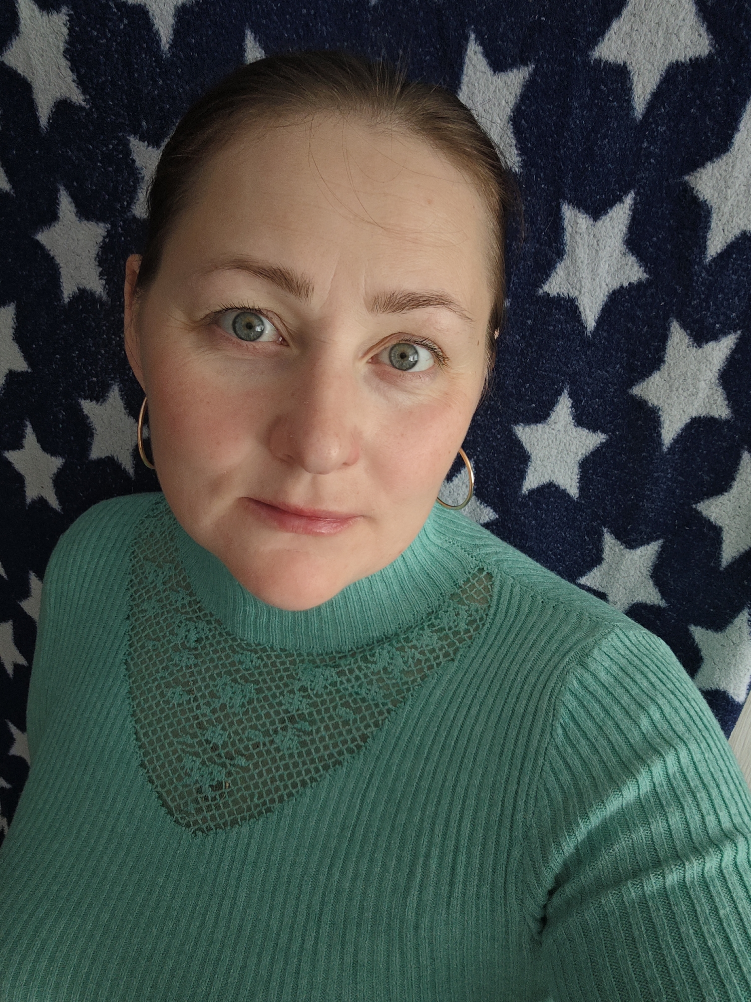 A close-up, selfie-style portrait features a woman looking directly at the camera. She has fair skin, wide-open green eyes, and light brown hair pulled back. Her complexion shows a slight flush on the cheeks, and minor lines are visible on her forehead. She is wearing a high-necked, ribbed sweater in a shade of teal or mint green, which has a V-shaped section of matching lace fabric across the upper chest. Gold hoop earrings adorn both ears. The background is a dark blue fabric patterned with numerous light-colored, five-pointed stars. The woman maintains a neutral to slightly contemplative expression. The setting appears to be indoors, potentially in Markovskii, Russia. No text is visible in the image.
