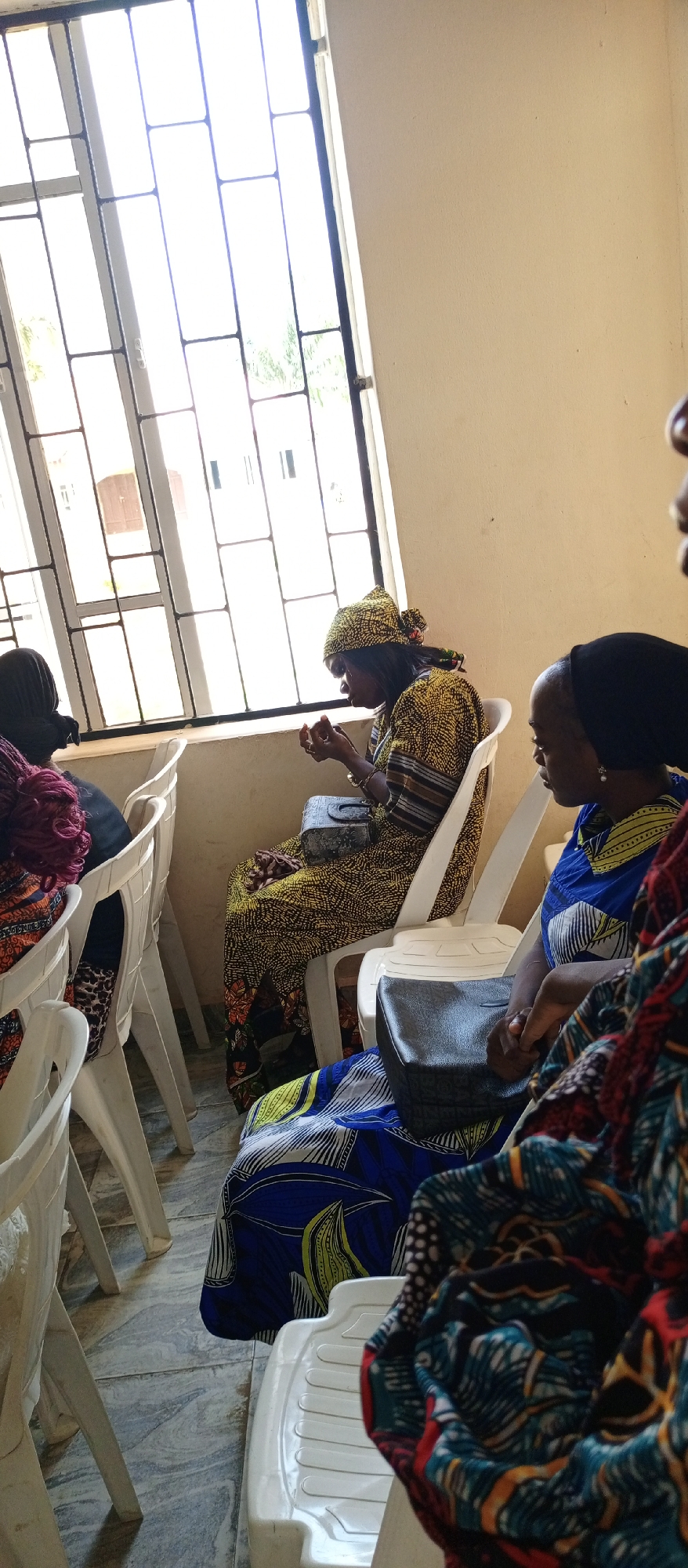 Inside a room in Keffi, Nigeria, several women are seated in white plastic chairs. One woman, clad in a yellow and black patterned dress and headscarf, sits near a window with barred panes and appears to be looking at something in her hands. Another woman, dressed in blue and yellow clothing and wearing a dark head covering, sits nearby. A third woman with red hair is partially visible to the left. A small black purse is on the lap of the woman in yellow. The room is lit by natural light from the window.