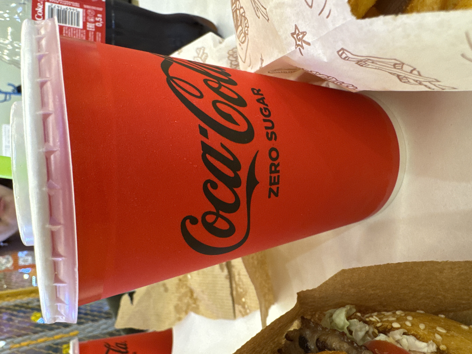 A close-up shot shows a red Coca-Cola Zero Sugar cup with a white lid and a green straw. The cup has the Coca-Cola logo printed in black. To the left, a portion of a burger wrapped in brown paper is visible. To the right, another wrapped food item with cartoonish drawings is present, and a dark bottle of Coca-Cola is partially visible in the background. The scene appears to be an indoor setting, possibly a fast-food restaurant, with blurred background elements suggesting a public space. The lighting is bright, and the focus is on the Coca-Cola cup. There is no discernible information in the image that would indicate a specific location in Oskemen, Kazakhstan.