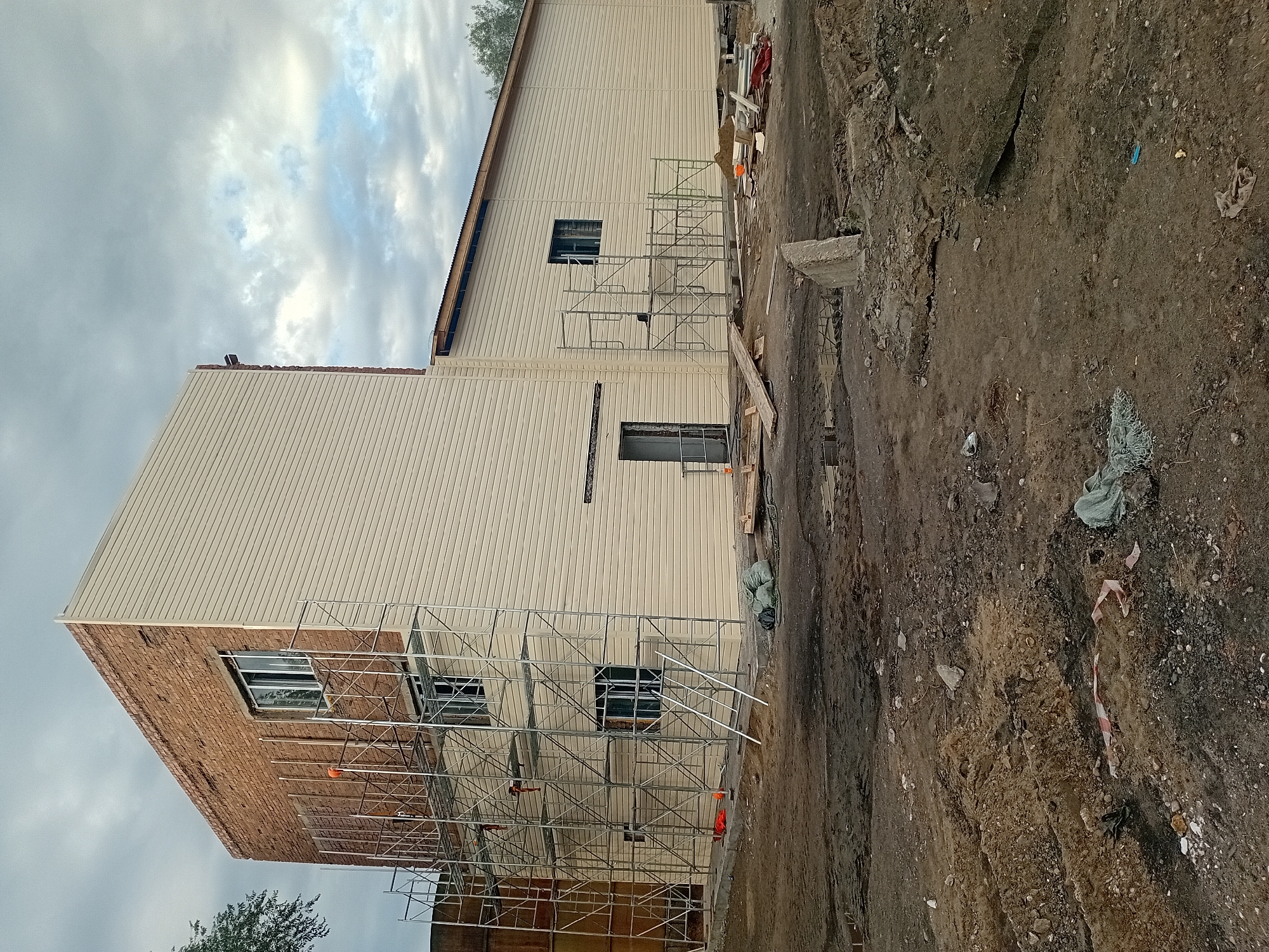 A construction site in Semey, Kazakhstan, features a partially completed building with scaffolding erected around a section of brickwork. The building has two distinct exterior finishes: exposed brick on the upper portion of one section and beige siding on the rest of the structure. Windows are visible on both the brick and sided sections. Several workers are present on the scaffolding, and various construction materials, including sacks of material and debris, are scattered on the muddy ground in front of the building. The sky above is overcast with clouds.