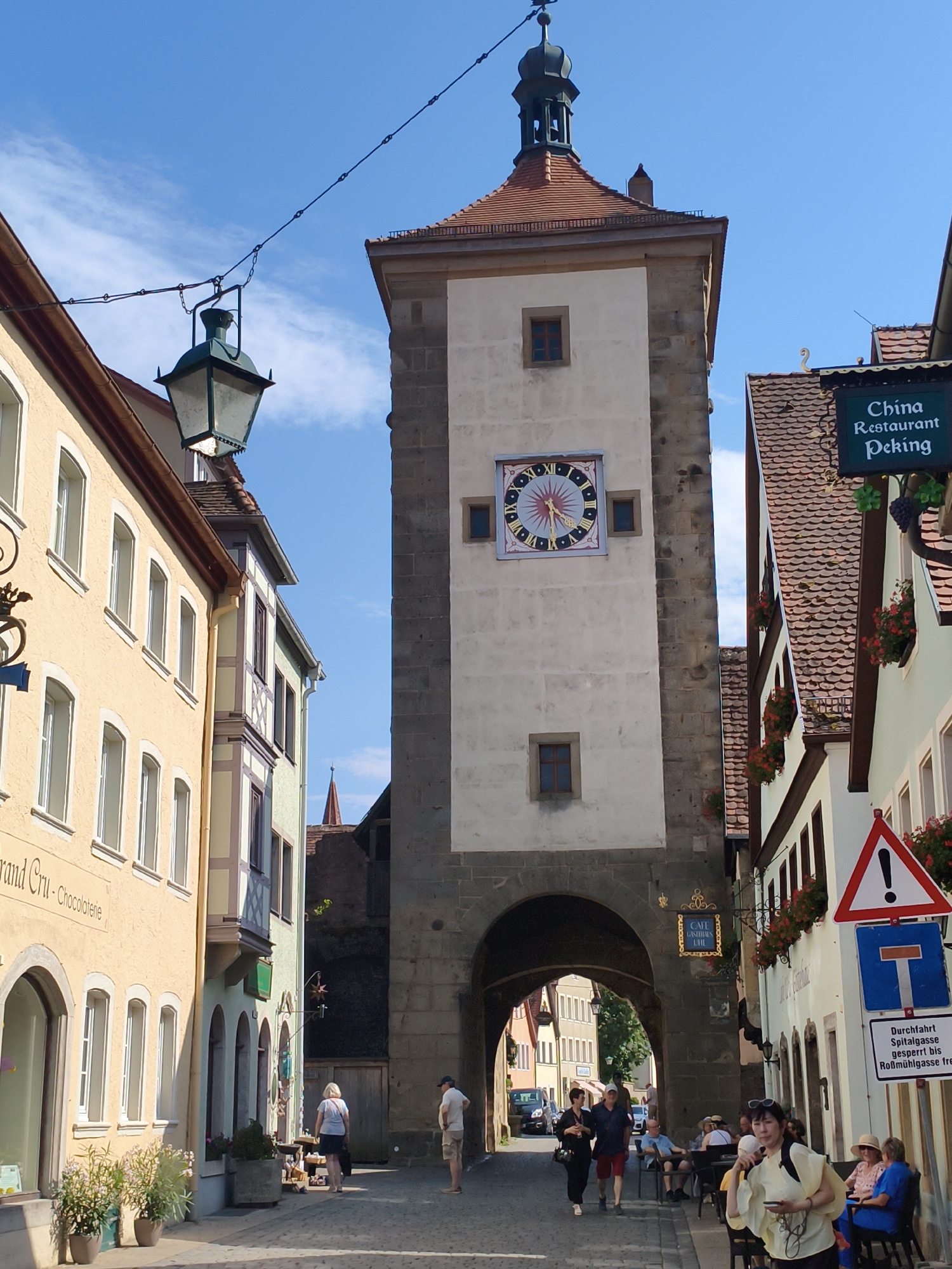 This image captures a street scene in Rothenburg ob der Tauber, Germany, focusing on the Plönlein intersection, a famous landmark. In the foreground, a cobblestone street leads towards a distinctive bifurcated building with a tower and an archway. The tower features a large clock face with Roman numerals and a weather vane at its apex. Several buildings line the street, displaying traditional German architecture with their tiled roofs, light-colored facades, and decorative window frames. One building on the right has a sign that reads "China Restaurant Peking." Various people are visible on the street, some walking, others seated at outdoor tables, suggesting a bustling, albeit relaxed, atmosphere. The sky is clear and blue with a few scattered clouds, indicating a pleasant day. The scene is filled with architectural details, signage, and hints of daily life in a historic European town.