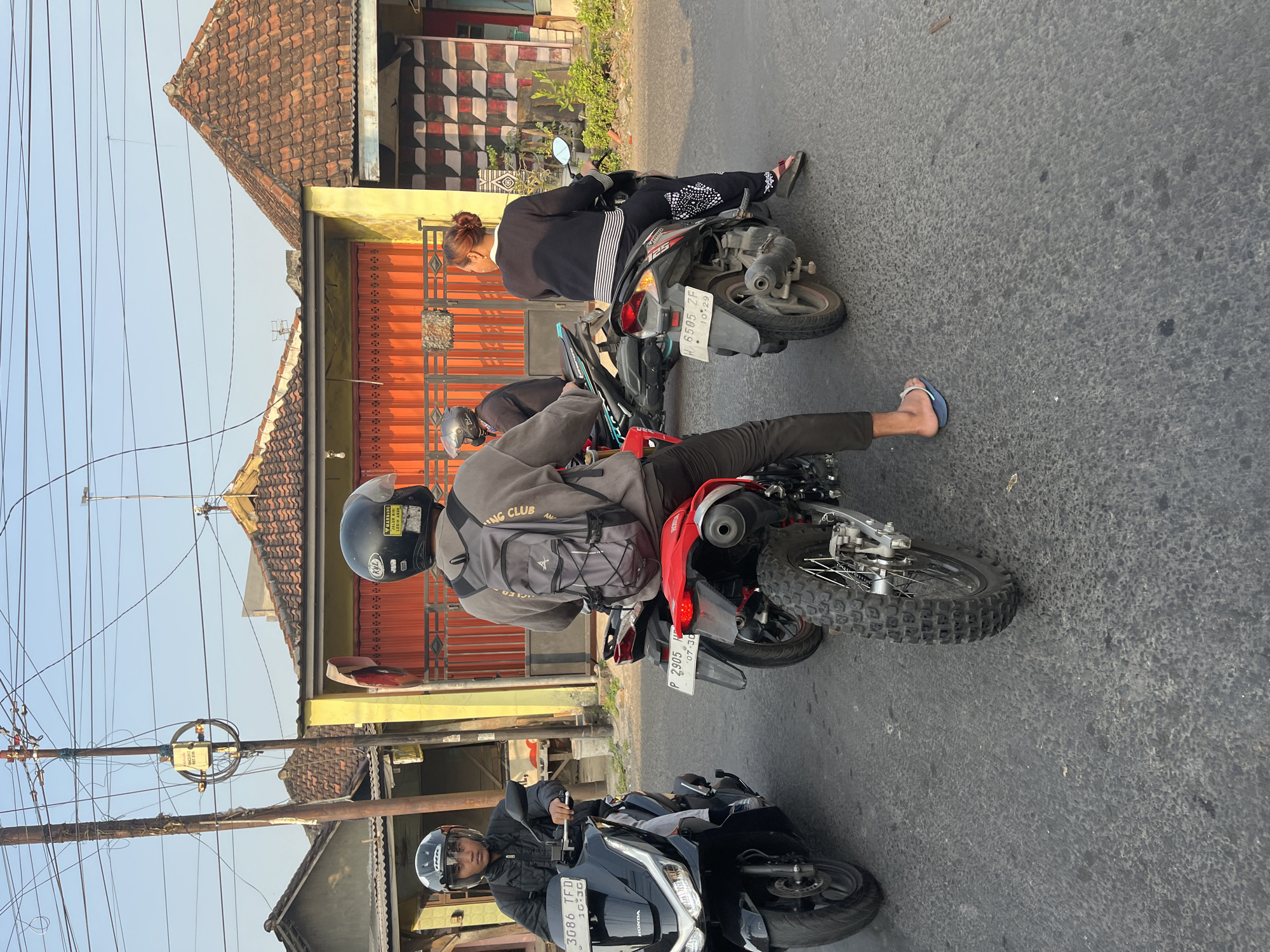In Sugih Waras, Indonesia, four individuals are observed on motorcycles on a paved road. The foreground features a black Honda scooter with a license plate reading "3086 TFD." To the right of the scooter is a red motorcycle with a license plate "P 2905 HF." Behind the red motorcycle is another scooter with a license plate "W 6505 ZF." The fourth motorcycle is partially visible, behind the scooter with the "W 6505 ZF" license plate.
The individuals are wearing helmets and are either operating their motorcycles or waiting. One person on the black scooter wears a black jacket. The person on the red motorcycle has a backpack and is facing away from the camera. The person on the "W 6505 ZF" scooter is a woman wearing dark clothing with white stripes and patterns. The fourth individual is only partially visible.
The environment suggests a residential area with buildings featuring tiled roofs and orange gates. Utility poles and wires are visible in the background. The lighting indicates it is daytime, likely late afternoon due to the warm tones. The road surface appears worn.