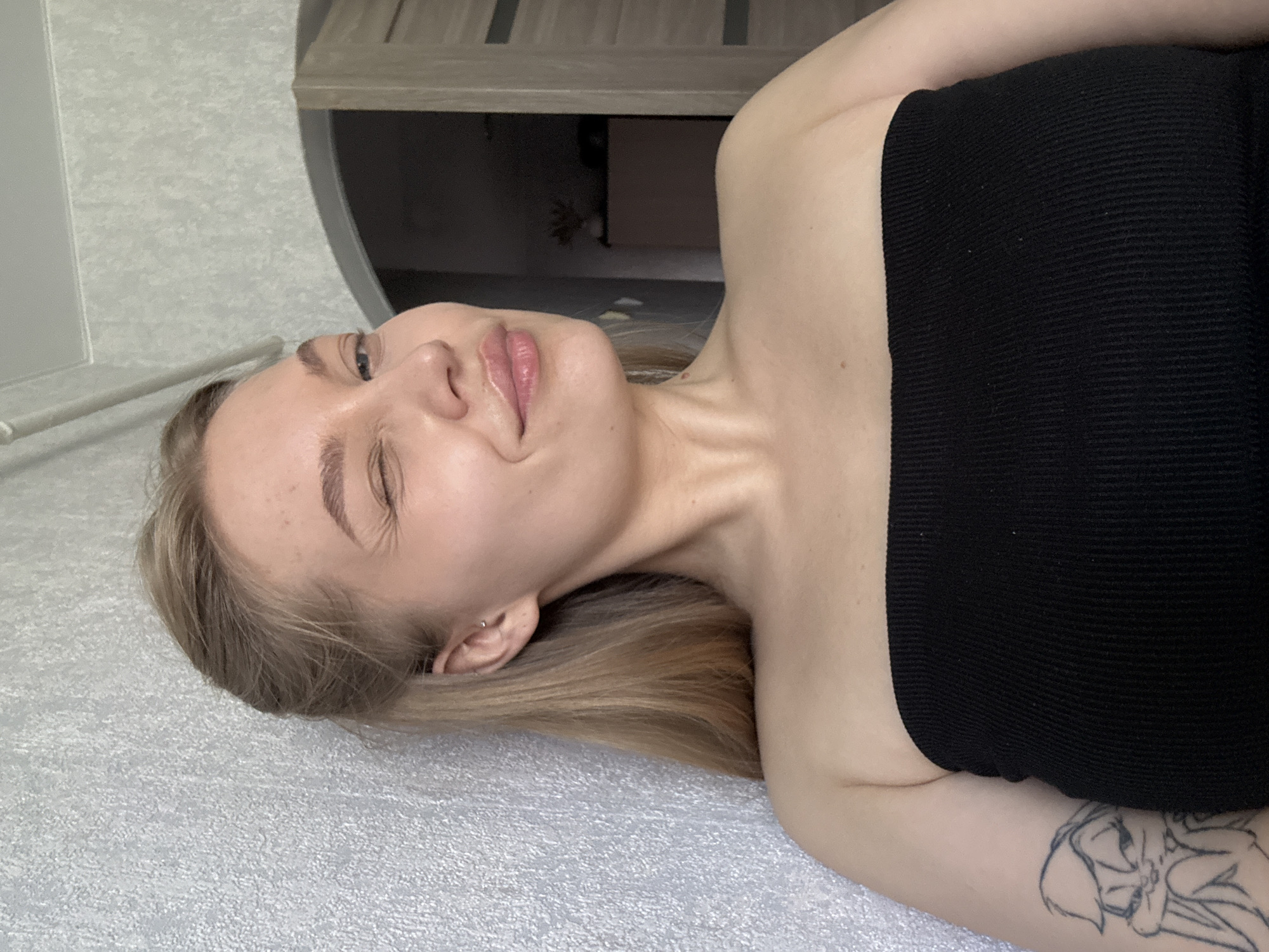 A fair-skinned individual lies on a textured, light grey surface, likely a bed or similar soft furnishing. She has long, blonde hair and is wearing a black, strapless top. Her face is turned slightly towards the camera with her eyes partially closed, suggesting a relaxed or sleepy state. There is a tattoo on her upper arm. The background includes a grey wall with a partially visible wooden shelf unit above. The image does not provide any information that can lead to the determination of the location in Chaikovsky, Russia.