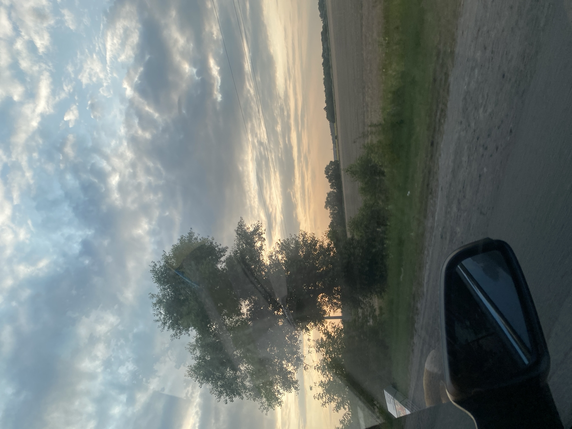 The image depicts a view from a vehicle, likely in or around the city of Pishchanka, Ukraine, during a sunset. The foreground features a car's side mirror reflecting the sky. Beyond the mirror, a dirt road runs alongside a grassy verge. A large tree with dense foliage dominates the mid-ground, silhouetted against the warm hues of the setting sun. Behind the tree, a field extends into the distance, with a line of trees marking the horizon. The sky is filled with dramatic clouds, ranging from light gray to darker tones, with streaks of orange and yellow indicating the sunset. Power lines are visible in the upper right portion of the frame. The overall impression is one of a rural landscape at dusk.