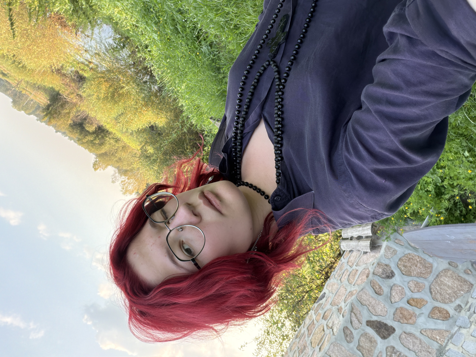 In Malyshevo, Russia, a young person with bright red hair and glasses is taking a selfie outdoors. They are wearing a dark blue collared shirt and multiple black beaded necklaces, one of which has a pendant. The background shows a stone structure to the left, lush green grass and vegetation, and a body of water. The sky is visible with scattered clouds.