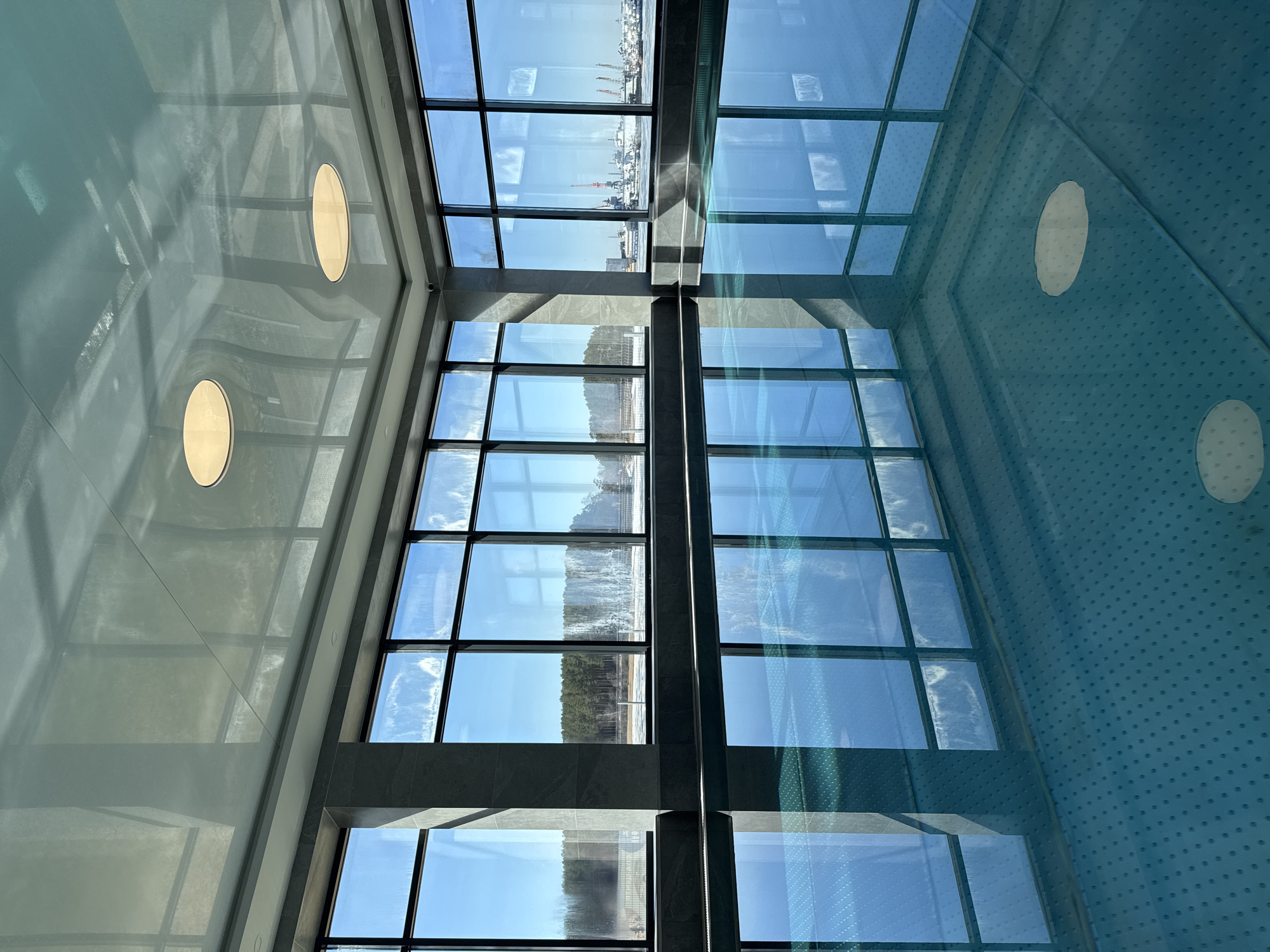 The image is an interior shot of a building in Chaikovsky, Russia. It is oriented at an angle, showcasing a ceiling with two round light fixtures on the left and a gridded window structure spanning the center. The windows reflect an outdoor scene. Through the windows, the landscape shows bare trees, and water and sky visible in the windows further in the frame, suggesting a coastline or riverside view. The right side of the image displays a blue surface with a grid of small dots and a blurry, circular light. The lighting appears bright and natural through the windows. The architectural details present clean lines and modern construction, suggesting a contemporary building design. No people are visible.