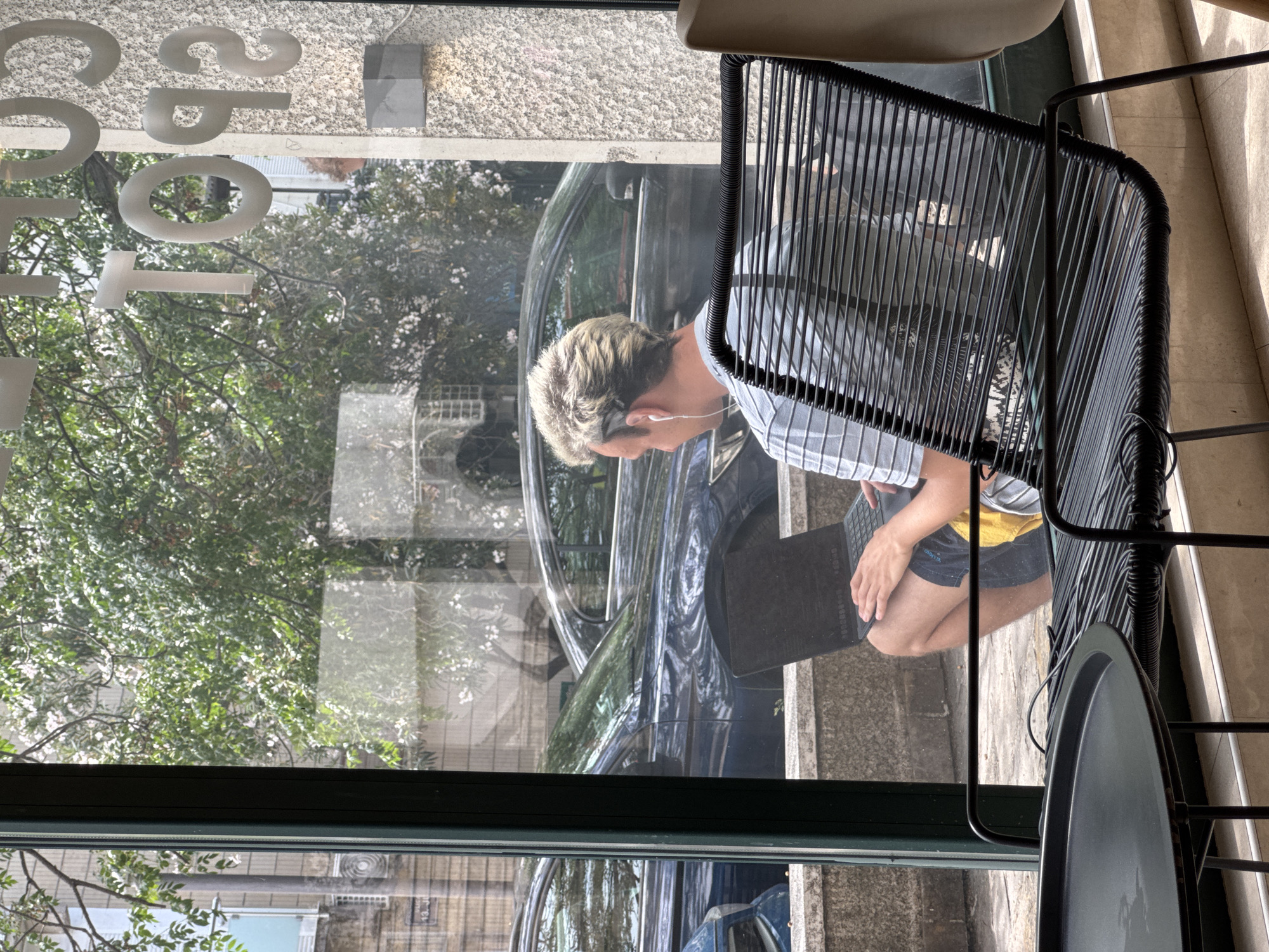 An image, taken from inside a establishment, shows a bright daytime scene through a large window overlooking a street in Budva, Montenegro. In the foreground, a person with short, light-colored hair is seated on a black woven chair, facing towards the left. They are wearing a light-colored striped shirt and dark shorts, with white earphones in their ears, and are actively engaged with a dark laptop resting on their lap. A small, round black table is partially visible to their right.
Through the clear window, the outdoor environment is visible, featuring lush green trees with scattered white blossoms, and a dark car parked on what appears to be a street or sidewalk. The bright sunlight creates clear reflections on the window surface, mirroring elements of the exterior. Mirrored text is visible on the upper part of the window, with individual characters resembling 'C', '0', 'T', 'T', 'E', 'P', '2', likely part of a business name or message, reversed from the perspective of an outdoor viewer. The person appears focused on their work or activity on the laptop, contributing to a sense of quiet concentration in a public setting.