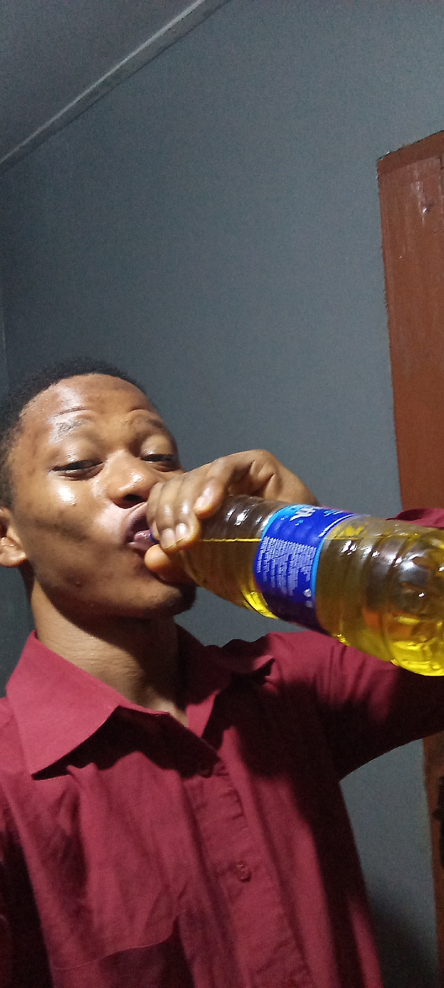 A person with dark complexion is drinking from a clear plastic bottle with a blue label. The bottle contains a yellow liquid. The person is wearing a red collared shirt. The background is a grayish-blue wall, with a brown wooden doorframe visible on the right side. The location is Unknown, Unknown.