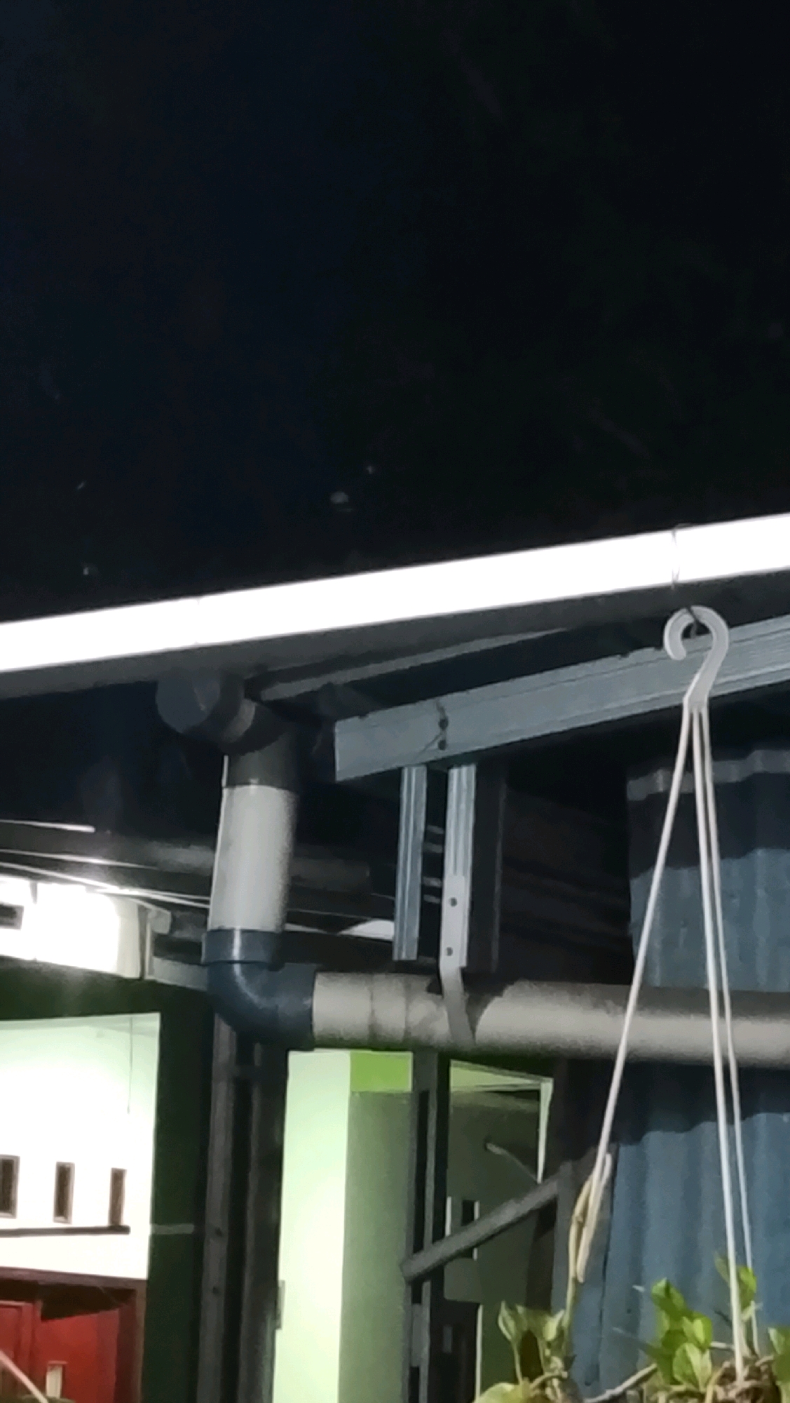The image shows an outdoor scene at night, likely in Ujungrusi, Indonesia, focusing on a section of a building's exterior. Prominently featured is a complex arrangement of PVC pipes, some white and some dark gray, connected by elbow joints. These pipes appear to be part of a drainage or utility system, running horizontally and vertically. Above the pipes, a white fluorescent light fixture is visible, casting a bright illumination on the scene. The background reveals corrugated metal siding, painted a dark blue or gray, and a portion of a building with light green walls and white windows with dark frames. A white plastic hook is suspended from an overhead structure, with thin white cords hanging from it, suggesting the presence of a hanging plant, some of which is visible in the lower right corner. The overall impression is of a functional, utilitarian area of a property, seen in low light conditions.