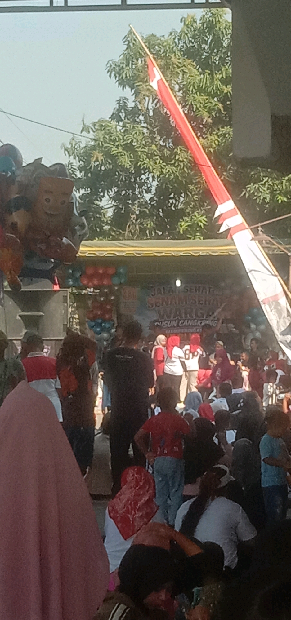 An outdoor event is taking place under a clear sky. A large red and white striped flag is prominently displayed, attached to a tall pole. In the background, a banner with Indonesian text is visible, reading "JALAN SEHAT SENAM SEHAT WARGA KUSUN CANGKOPAN MUNGGING". This suggests the event is a healthy walk and exercise activity for the residents of Kusun Cangkopan in Mungging, Temuwulan, Indonesia.
A stage area is set up, decorated with balloons in red and blue. Several people are gathered on and around the stage, with some appearing to be performers or speakers. A group of people, likely participants, are standing in front of the stage. Children are visible among the crowd.
To the left, a cluster of large, inflatable cartoon-like figures are present, possibly part of the event's decoration or entertainment. The overall scene depicts a community gathering likely focused on health and well-being. The presence of the Indonesian flag and the text on the banner confirm the location and context of the event.