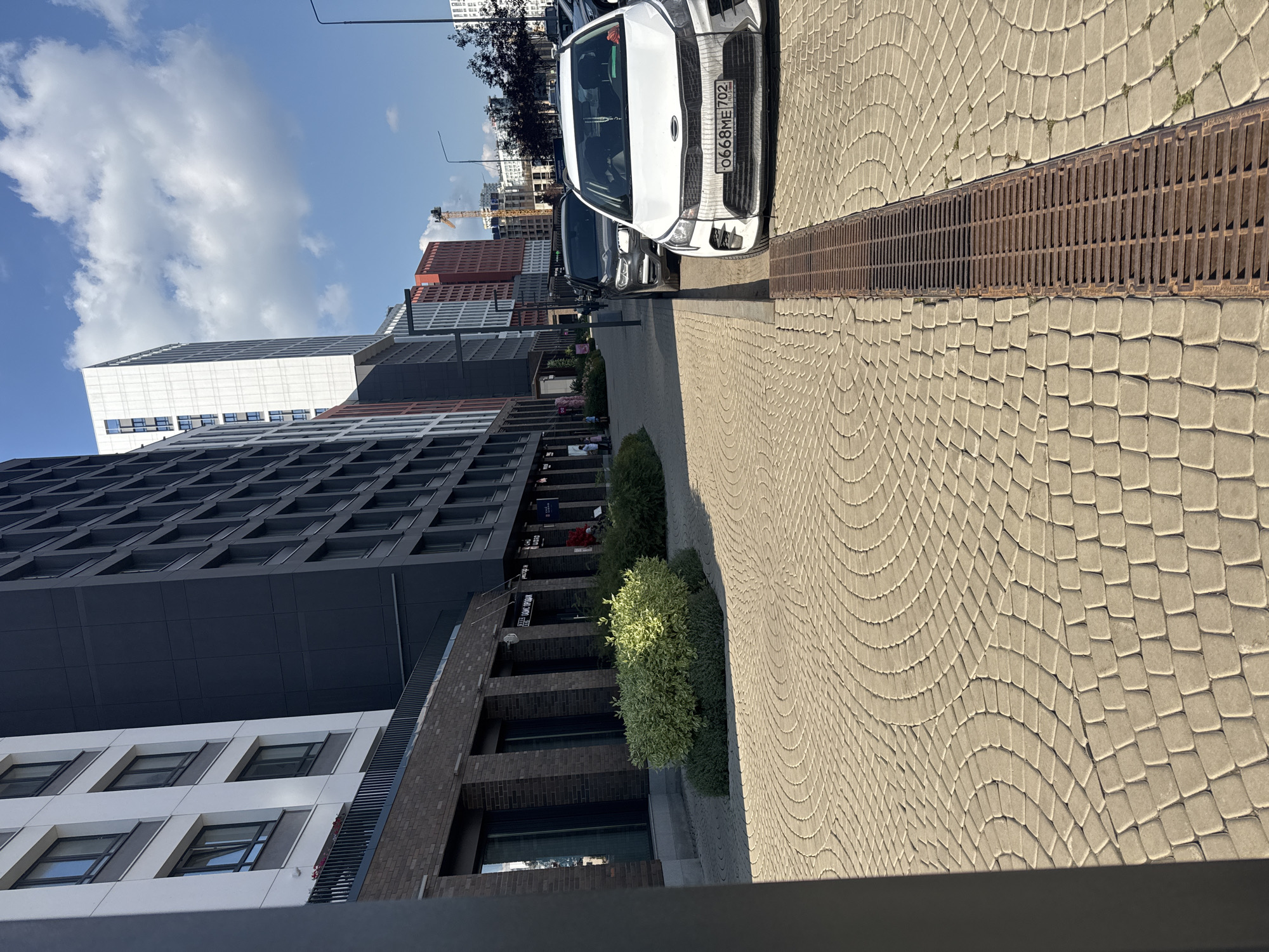 The image captures a sunny day in Ufa, Russia, with a street scene dominated by modern architecture. In the foreground, a cobblestone pathway features a drainage grate. Several cars are parked along the street, including a white Kia Rio with a Russian license plate that reads "0668ME 702". Behind the cars, a row of contemporary buildings rises, characterized by their dark facades and numerous windows. Further in the background, other buildings of varying designs and heights are visible, along with a construction crane, suggesting ongoing urban development. Greenery in the form of bushes and trees adds a touch of nature to the urban landscape. The sky is blue with scattered white clouds.