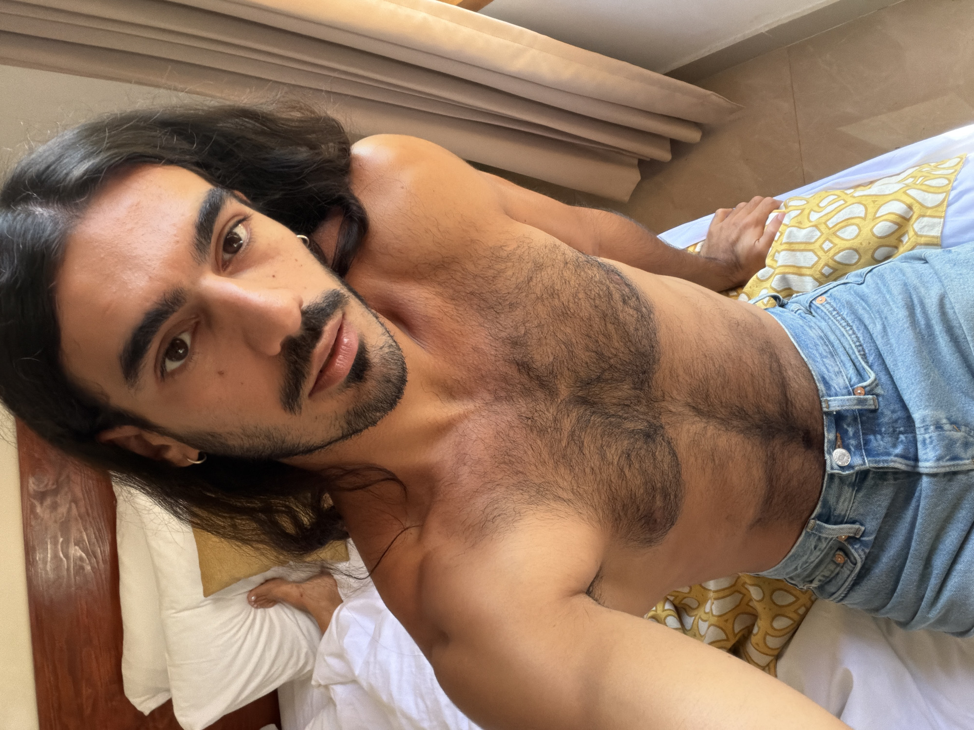 A person with long, dark hair, a dark mustache, and a beard lies on a bed. The person is shirtless and wearing light blue denim jeans. The bed has white sheets and a yellow-and-white patterned pillow. In the background, there is a window with beige curtains and a beige tiled floor. The picture was likely taken in an interior setting, possibly a hotel or residence in North Kuta, Indonesia.