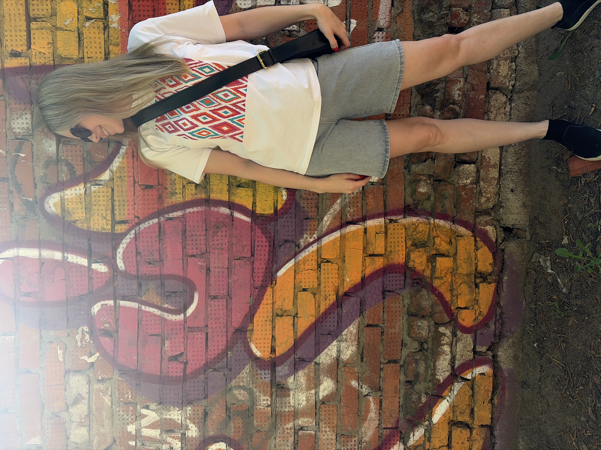 The image shows a woman lying horizontally against a brick wall in an outdoor setting. The woman is wearing sunglasses, a white T-shirt with a patterned design, denim shorts, and black shoes with white soles. She has long blonde hair and a black bag slung across her chest. The brick wall features colorful graffiti in shades of red, orange, and white. The right side of the image shows a patch of dirt and some vegetation. The photograph appears to be taken from a top-down perspective. It is not possible to confirm the location as Chaikovsky, Russia without additional information.