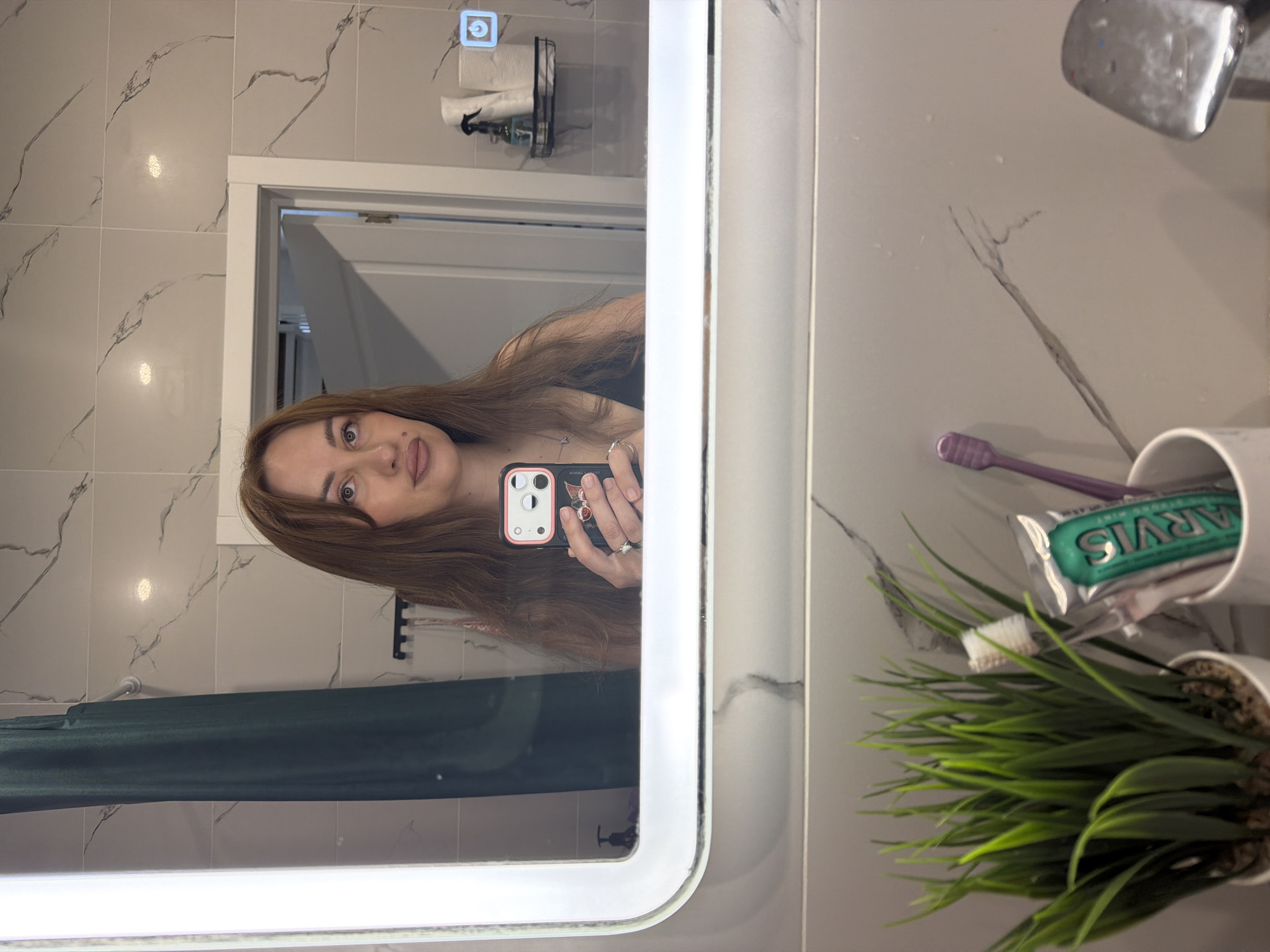 A woman with long, reddish-brown hair and fair skin is taking a mirror selfie in a bathroom. She is wearing a dark top and is holding a smartphone with a pink cherry-themed case. She has rings on her fingers and a necklace. The bathroom has marble-patterned tiles on the walls and a large, illuminated mirror. In the reflection, a dark green shower curtain is visible. On a shelf in the background, there are toiletries including a spray bottle and rolls of toilet paper. In the foreground, there is a white pot with artificial green grass, a toothbrush, and a tube of Marvis toothpaste. Another purple toothbrush is in a white cup to the right of the toothpaste. The overall scene suggests a personal grooming area.