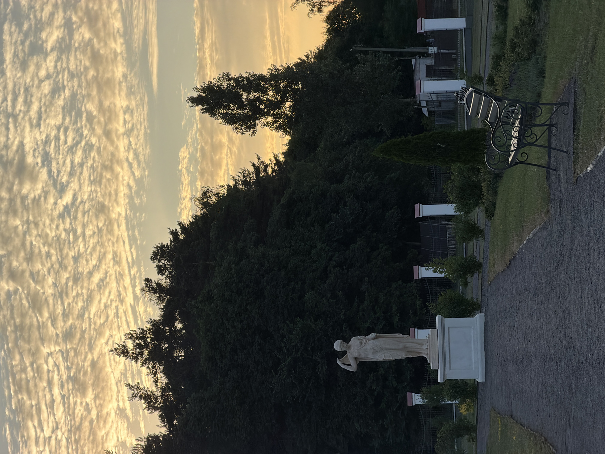 An outdoor scene depicts a statue of a classical female figure, likely a deity or muse, positioned centrally on a white pedestal. The statue is made of light-colored stone and is draped in flowing robes. Behind the statue, a dense line of dark green trees forms a natural backdrop. The sky above is filled with dramatic, layered clouds illuminated by the warm hues of either sunrise or sunset, casting a soft, golden light.
In the foreground, a dark gravel path leads towards the statue. To the right of the path, a dark metal bench with intricate scrollwork is placed on a patch of green grass. This bench faces towards the statue, suggesting a contemplative or viewing area.
Further in the background, a white fence with decorative pillars and dark metal fencing encloses a manicured garden area with small, neatly trimmed bushes. A tall, slender evergreen tree stands prominently to the right of the statue, contrasting with the broader foliage of the trees behind. The overall impression is one of a serene, possibly memorial or decorative garden space, enhanced by the dramatic sky.