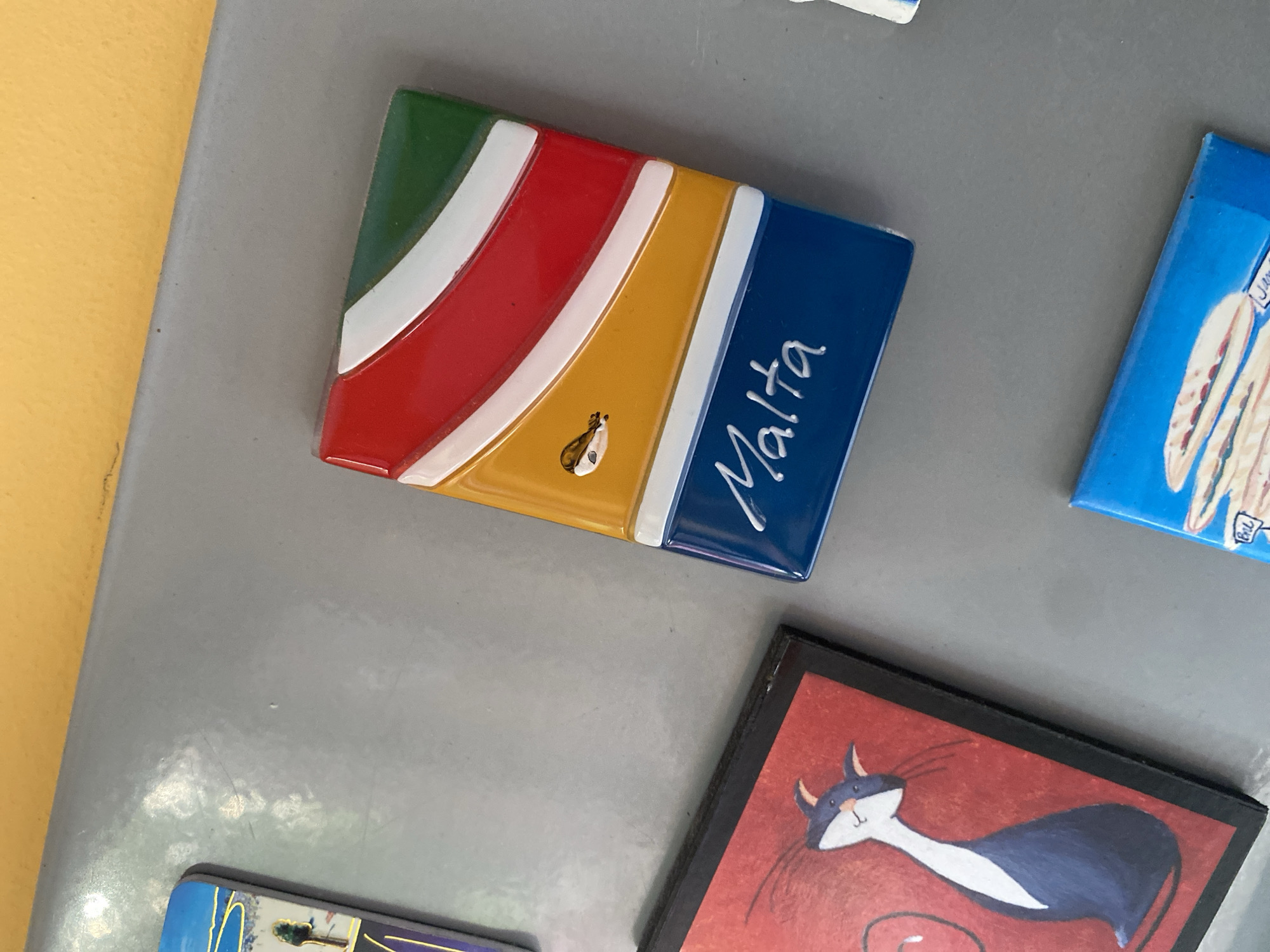 A close-up, top-down shot shows several refrigerator magnets attached to a gray metallic surface, likely a refrigerator door. The magnets display various designs. Prominently featured is a magnet depicting the flag of Malta, with its characteristic white and red design and a yellow section. The word "Malta" is written in white script on the lower blue portion of this magnet. To the left, a magnet with a red background and a stylized black and white cat is visible. Another magnet, partially visible at the top left, appears to have a landscape scene with a tree. At the bottom of the frame, a portion of a blue magnet shows a graphic of what looks like sandwiches. The background above the metallic surface is a solid, vibrant yellow. The scene is devoid of people and any discernible signs of the city of Wrocław, Poland.