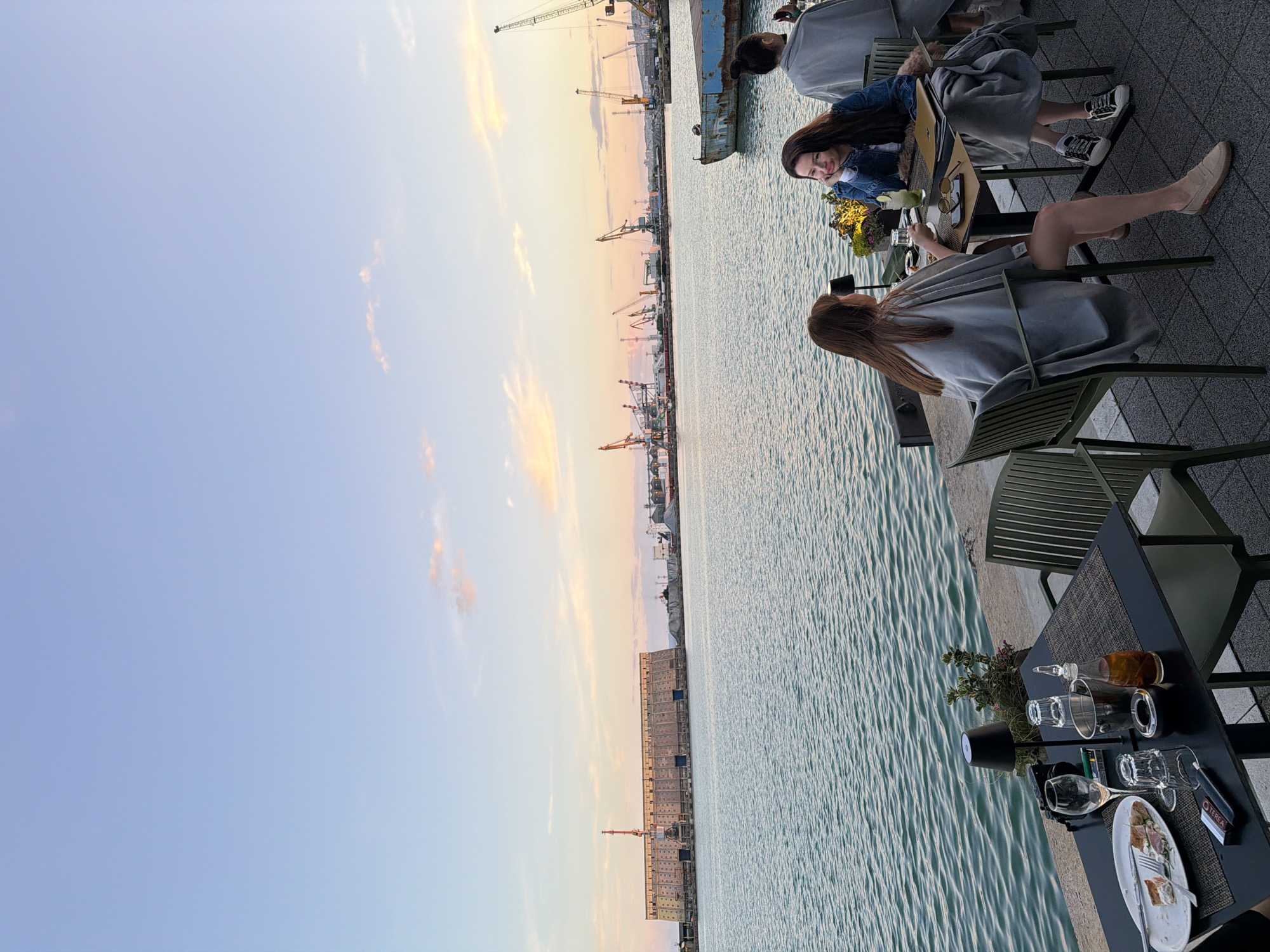 This is an eye-level, outdoor, medium shot of a waterfront dining scene in Burgas, Bulgaria, during dusk. The sky is a gradient of light blue to pale orange, with a few wispy clouds. The water is a calm, rippling turquoise.
In the foreground, a black table is set for dining, with two place settings. One place setting includes a plate with a partial meal, cutlery, a wine glass, and two cigarette packs. Another place setting has two glasses, an ashtray, and a small bottle of oil. A small black lamp and a potted plant are also on the table. To the right of the table, two women are seated. The woman on the left, facing away from the camera, is wearing a grey shawl and has long brown hair. The woman on the right, facing the camera and leaning on her hand, is wearing a denim jacket and has long dark hair. She is holding a drink with a straw. Behind them, another woman in a grey shawl is seated, facing away from the camera.
In the middle ground, the turquoise water extends to a concrete embankment. Beyond the water, there is a large industrial waterfront with numerous cranes, ships, and large buildings. Notably, a long, imposing building structure with many windows is visible on the left side of the harbor.
The overall atmosphere is serene and relaxed, with the soft evening light casting a gentle glow over the scene.