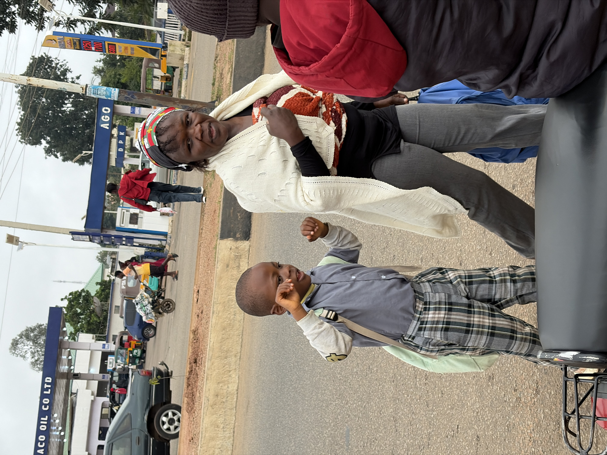 An overcast day in Jos, Nigeria, shows a woman in the foreground with a white shawl draped over her shoulders and a colorful patterned garment visible underneath. She wears grey trousers. To her left, a young boy in a grey collared shirt and plaid trousers, carrying a light green bag, appears to be rubbing his eye. Behind them, several other people are walking or standing near a gas station. The gas station features blue awnings with white lettering that reads "AGO OIL CO. LTD" and "AGO." A price board with red and white digits is visible on the right. The environment includes a paved road, a concrete barrier, and various vehicles, including cars, a tricycle, and what appears to be a motorcycle in the very foreground. The overall scene suggests a busy street in an urban setting.
