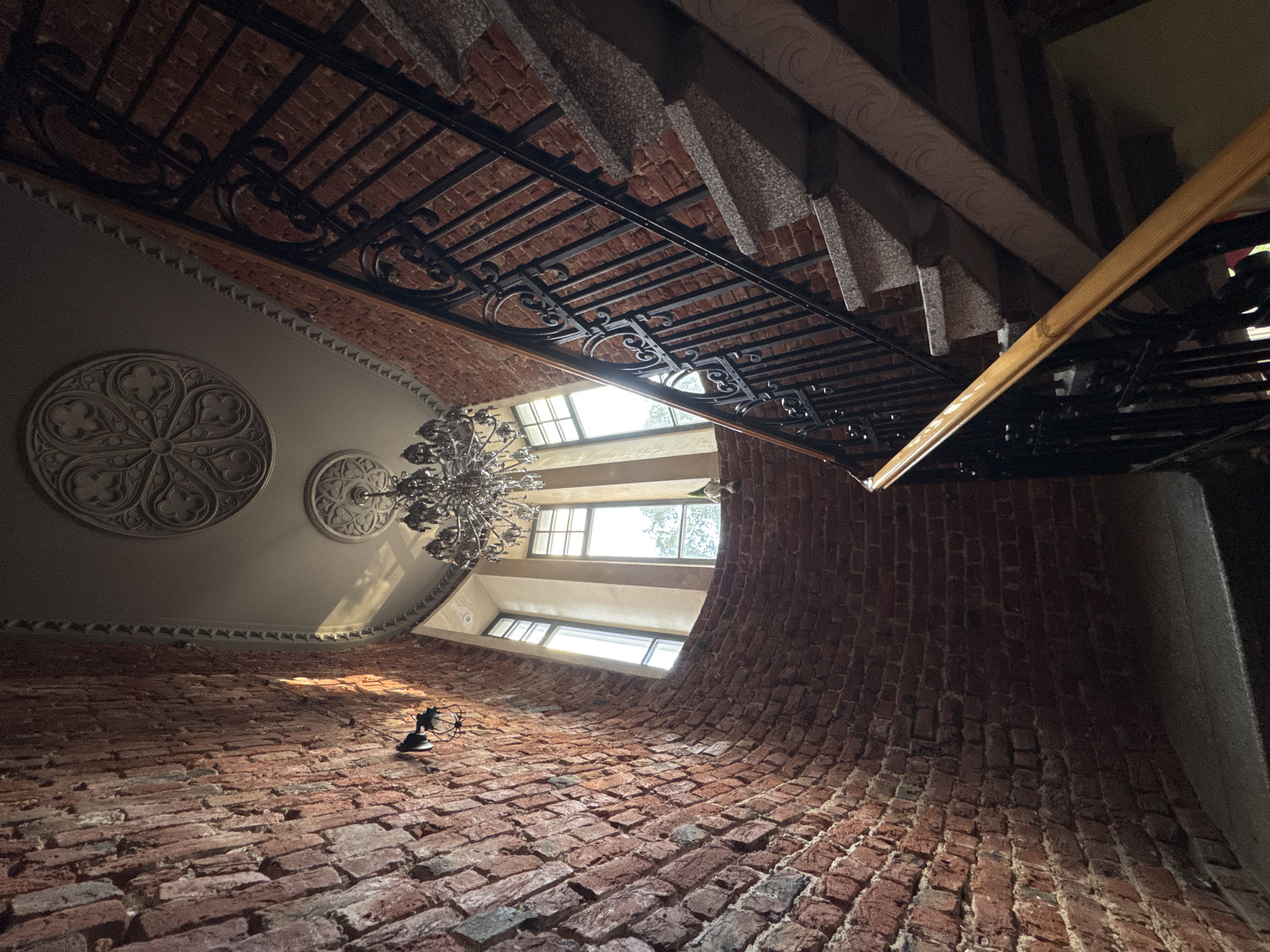 This media file presents a low-angle, fisheye perspective of an interior architectural space, likely a grand staircase or atrium in Saint Petersburg, Russia. The primary subjects are the textured brickwork of a curved wall that dominates the lower half of the frame, and the ornate ironwork of a railing and ceiling structure above. A grand chandelier hangs from the ceiling, and decorative circular reliefs are visible on the plastered upper walls.
The scene appears to be illuminated by natural light filtering through a series of windows set high in the curved wall. The time of day is not definitively discernible, but the quality of light suggests it could be daytime. The weather outside is not apparent.
Notable details include the rough texture and varied hues of the exposed red bricks, suggesting an older building. The intricate design of the black iron railing and ceiling elements adds a touch of historical elegance, contrasting with the raw brick. The decorative ceiling rosettes are classic architectural ornamentation. A small, dark object, possibly a lamp or a decorative fixture, is mounted on the brick wall.
The perspective creates a sense of depth and scale, emphasizing the architectural features. The absence of people or active events makes the focus entirely on the static, yet dynamic, architectural elements. There is no visible text in the image. The overall impression is one of historical grandeur and perhaps a slightly dramatic or imposing atmosphere due to the extreme perspective.