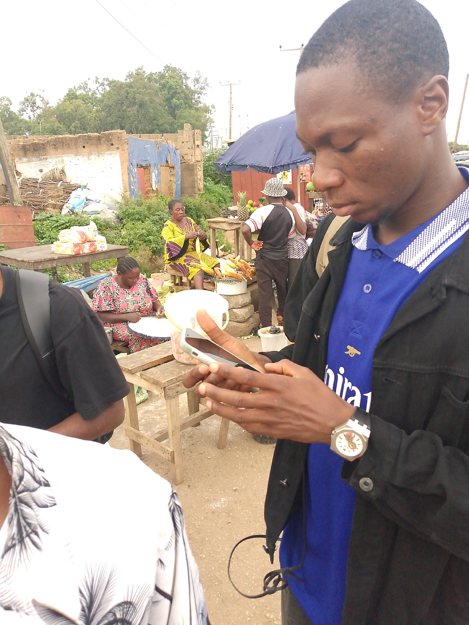 In Bukuru, Nigeria, a young man in a black jacket over a blue collared shirt is engrossed in his phone. He wears a wristwatch on his left arm. Behind him, a market scene unfolds. Two women are visible. One, in a yellow and purple patterned dress, sits on a stool at a stall, seemingly preparing food. Another woman, in a pink and black patterned dress, is seated at a wooden table, also working with food items laid out on a white plate. A table laden with packaged bread and other goods is to the left, and a display of corn on the cob and pineapples suggests fresh produce is being sold. A blue tarp forms an awning over part of the market. The setting appears to be an outdoor market, with some dilapidated structures in the background and trees visible above. The ground is dirt and gravel. The overall impression is of a typical day at a Nigerian market.