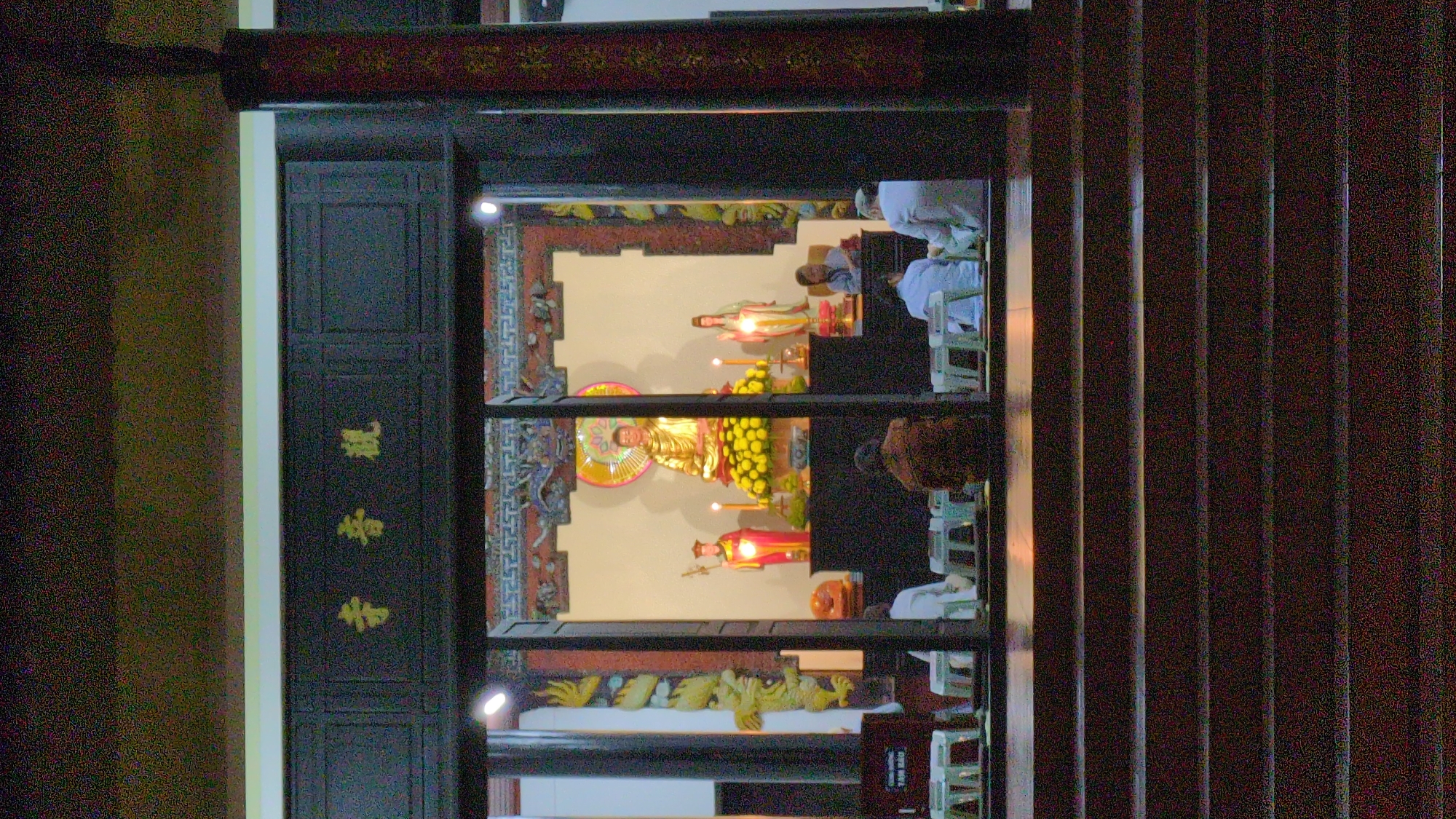 The image captures a view into a Buddhist temple located in Da Lat, Vietnam. Three levels of the temple's interior are visible through a doorway. Each level displays Buddha statues adorned with flowers and lights. People in white are observing. The scene is framed by ornate dark wood structures with gold calligraphy, likely part of the doorway or window. The side of a wooden structure is also visible.