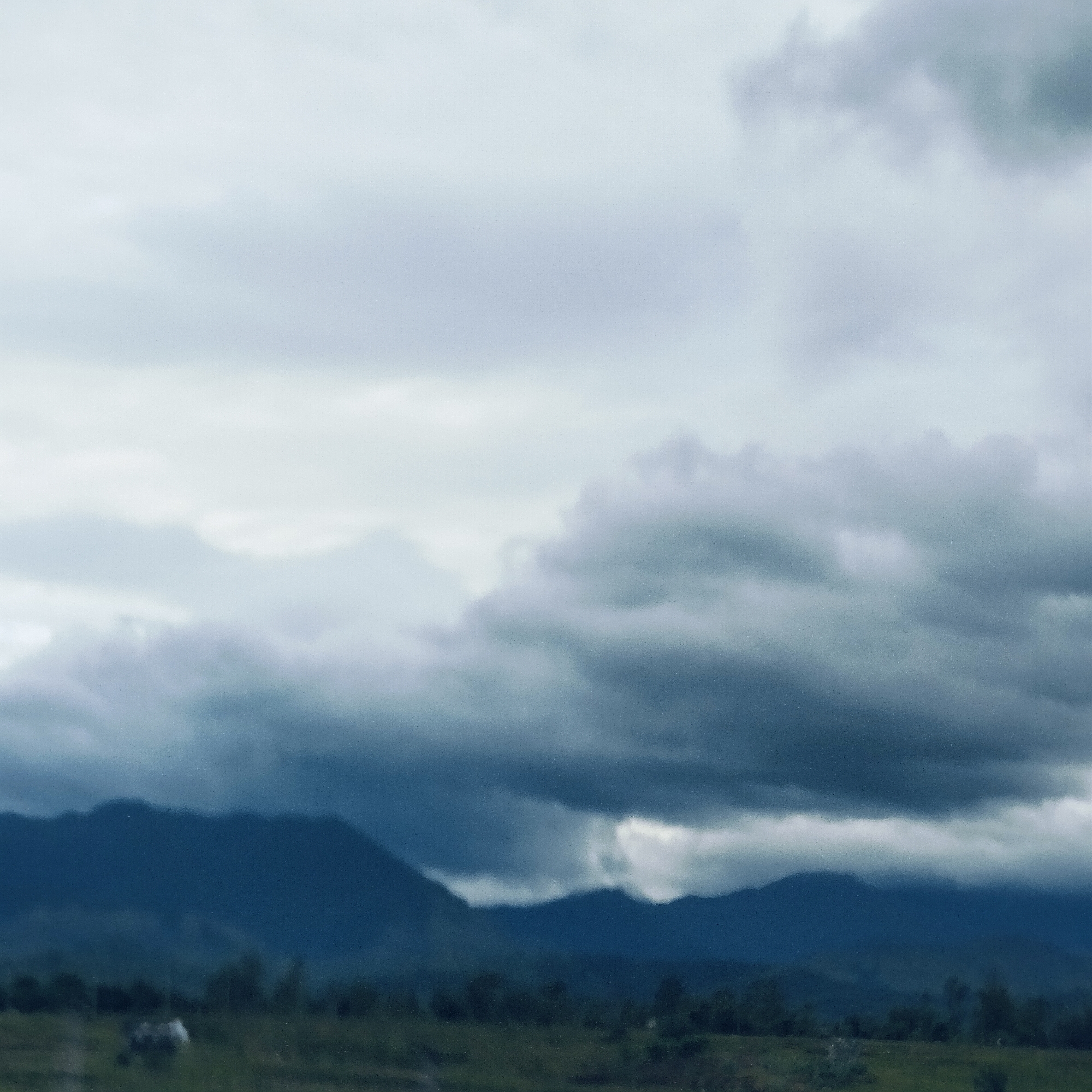 The image presents a wide, overcast landscape, likely captured during the day in Keureuweungkrueng, Indonesia. The scene is dominated by a dramatic sky filled with thick, textured clouds ranging in color from pale grey to deep, moody blue-grey, suggesting heavy cloud cover or an approaching storm. These clouds occupy the majority of the frame, creating a vast and somewhat somber atmosphere.
In the mid-ground, a range of dark mountains rises, their undulating peaks appearing as deep blue-grey silhouettes against the cloudy sky. Their forms are partially softened by atmospheric haze and the diffuse light, seamlessly blending with the darker clouds in places.
The foreground consists of a blurry, dark green expanse, likely a field or open land. A faint, light-colored shape is barely discernible in this area, possibly an animal, though its details are too indistinct to confirm. The overall color palette is composed primarily of cool tones, with blues, grays, and dark greens lending a serene yet powerful feel to the natural vista. There is no visible text in the media file.