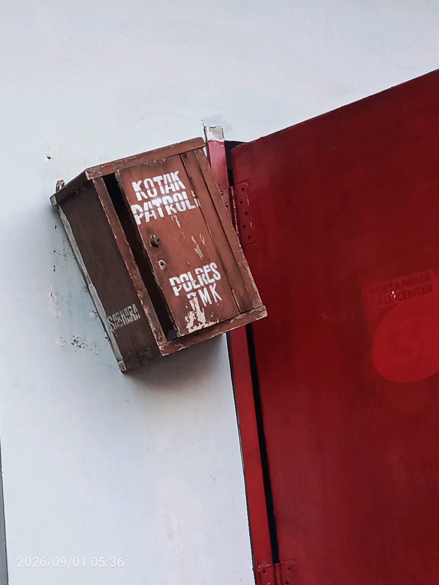 A medium shot captures a distressed, wooden patrol box mounted on a white wall to the left, adjacent to a bright red metal door on the right. The box, tilted at an angle, displays white stenciled text that reads "KOTAK PATROLI" at the top and "POLRES 7MK" below. A faded, illegible inscription in a similar stencil is visible on the left side of the box. The door on the right features faint red lettering that appears to say "PERTAMINA CALL CENTER". The scene is illuminated by natural light, casting subtle shadows. The timestamp "2026/09/01 05:36" is present in the bottom left corner of the image. The background is a plain white wall with some visible marks and scuffs. The overall environment suggests an outdoor or semi-outdoor location, possibly in an urban setting given the presence of the call center text. The exact location in the city of Gladak Anyar, Indonesia, is not explicitly visible but inferred from the surrounding context of the request.