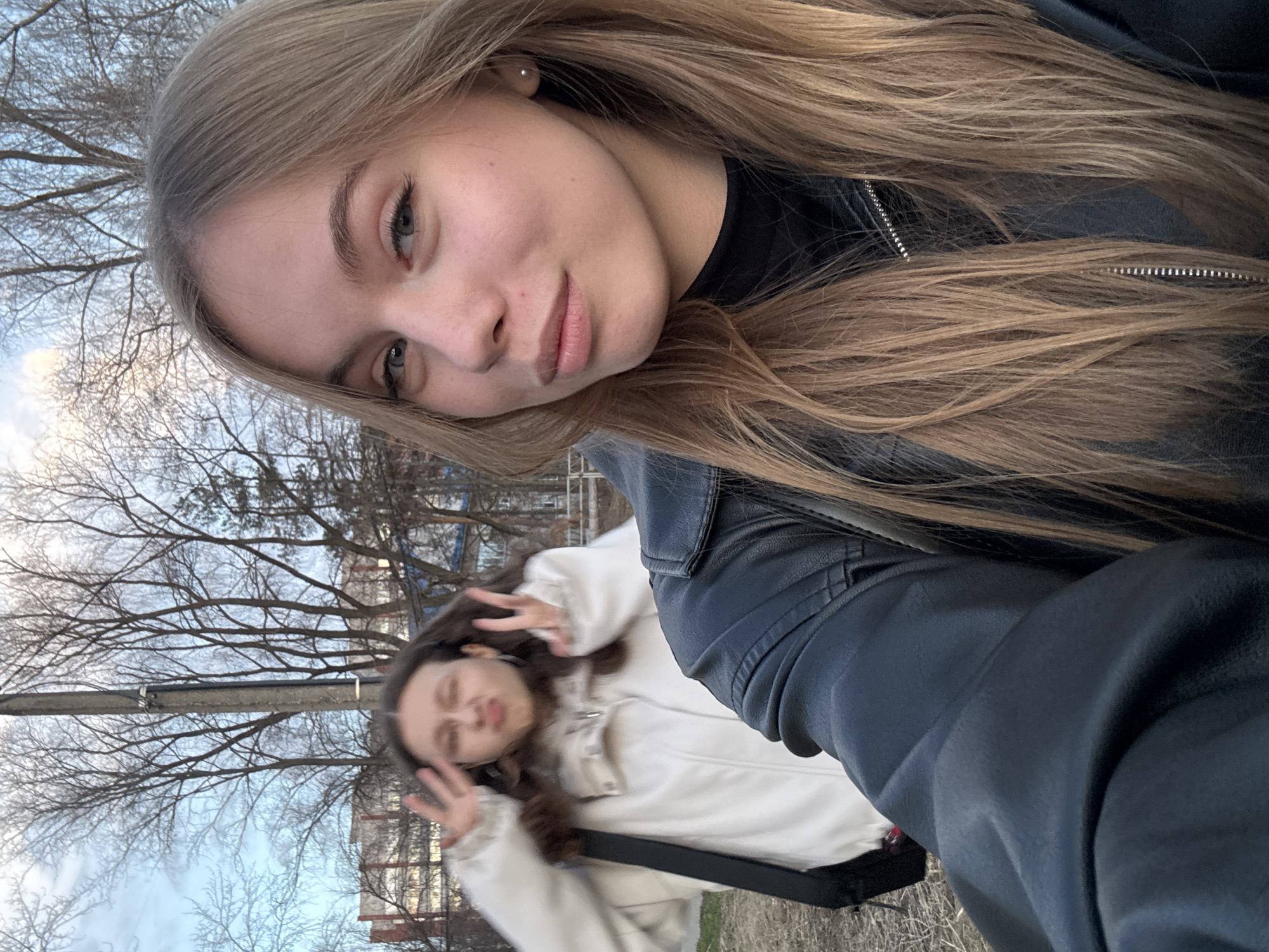 A tilted selfie shows two young women. The woman in the foreground is closer to the camera, wearing a black leather jacket and a black top. She has fair skin, blond hair, and is looking at the camera with a slight smile. Behind her and slightly out of focus is another woman with darker hair, wearing a white jacket and making a peace sign with both hands. The background includes bare trees, power lines, some buildings, and a glimpse of the sky. The location appears to be outside, possibly in a park or urban area in Chaikovsky, Russia.