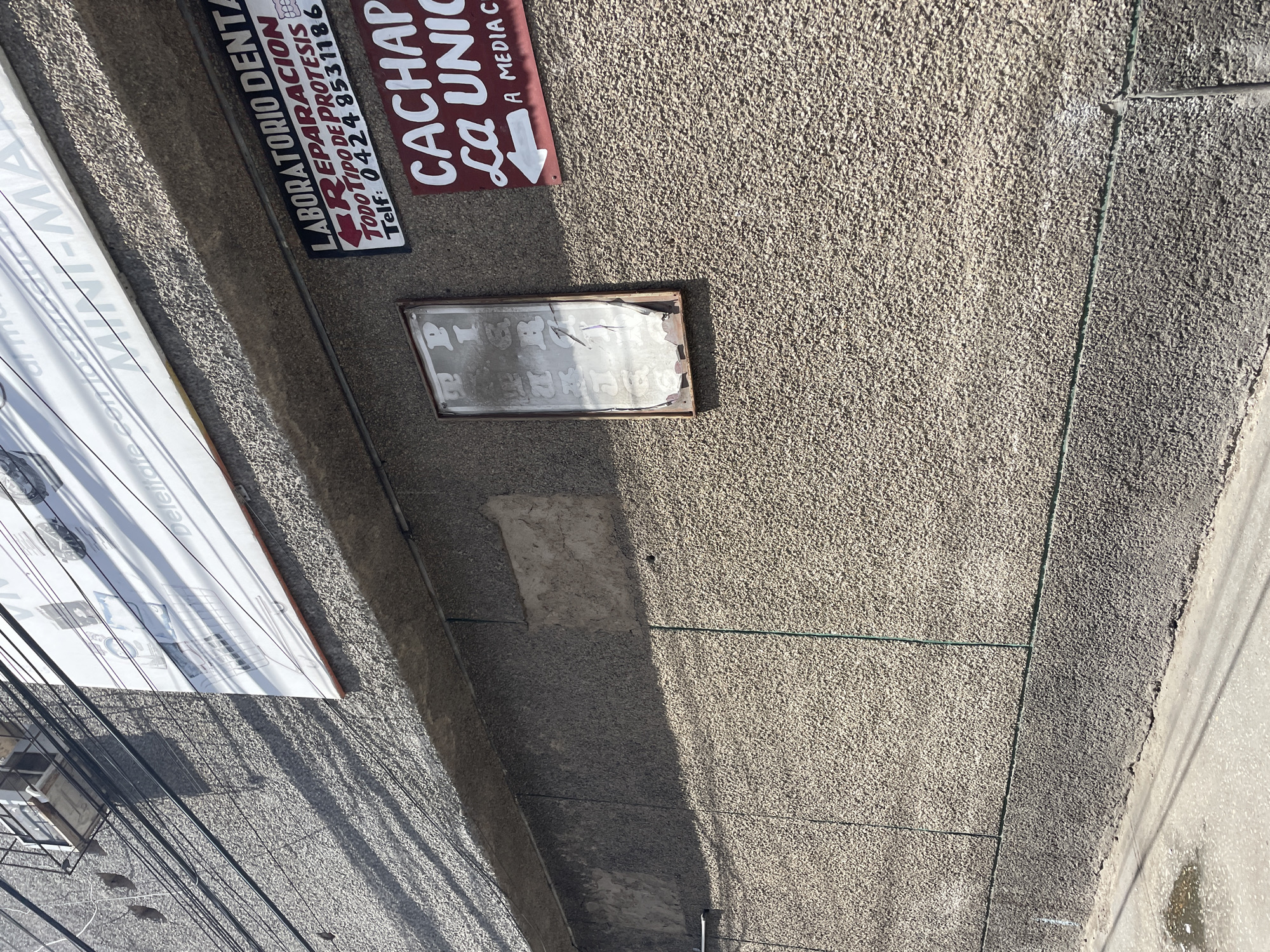 The image shows a textured concrete wall with several signs and an air conditioning unit visible. At the top left, an outdoor air conditioning unit is mounted on a black metal bracket. Below it, a large white sign with red and black text is partially visible, with the words "Deleit" and "MINI-MA" discernible. To the right of this sign, a dark rectangular sign reads "LABORATORIO DENTAL REPARACION TODO TIPO DE PROTESIS TelF: 0424 8531186". Below this, another dark red rectangular sign with white text states "CACHAPA LA UNICA A MEDIA C". On the wall, a vertical, distressed, framed sign with white lettering is also visible. The wall is streaked with shadows cast by overhead wires. The ground in the foreground shows a patch of wet concrete and more overhead wiring. The scene appears to be an outdoor urban environment.