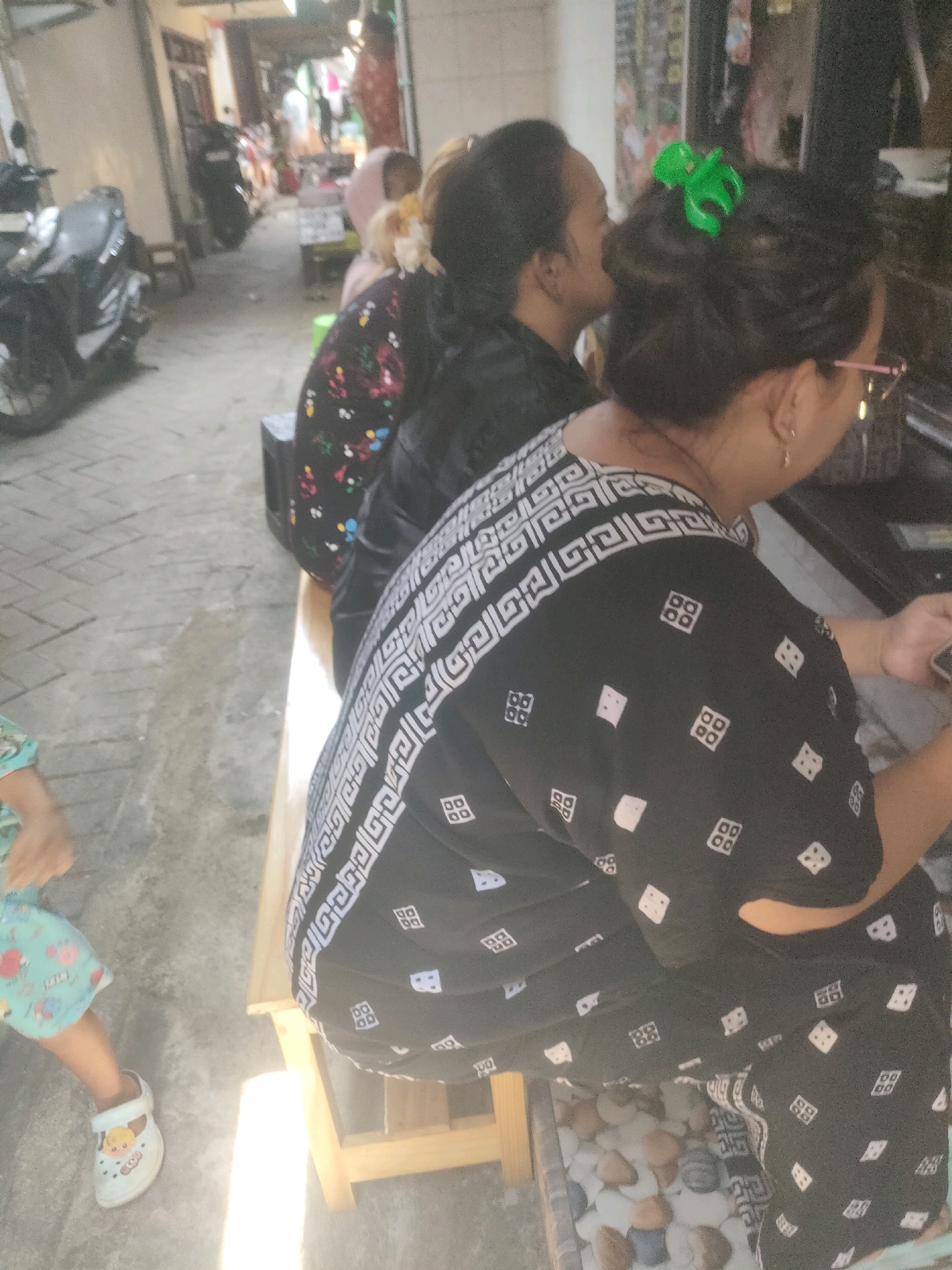 The image shows an outdoor scene in Surabaya, Indonesia. In the foreground, a woman wearing a black garment with a white geometric pattern is seated on a wooden bench. Her hair is pulled back and held by a bright green hair clip. Next to her, another woman in a black top with a multi-colored floral print is also seated. To the left, a child is visible, wearing a light blue patterned skirt and white clogs. The background depicts a narrow street or alley lined with buildings, with a motorcycle parked on the left side. The ground is paved with cobblestones. The lighting suggests it is daytime, with sunlight casting shadows.