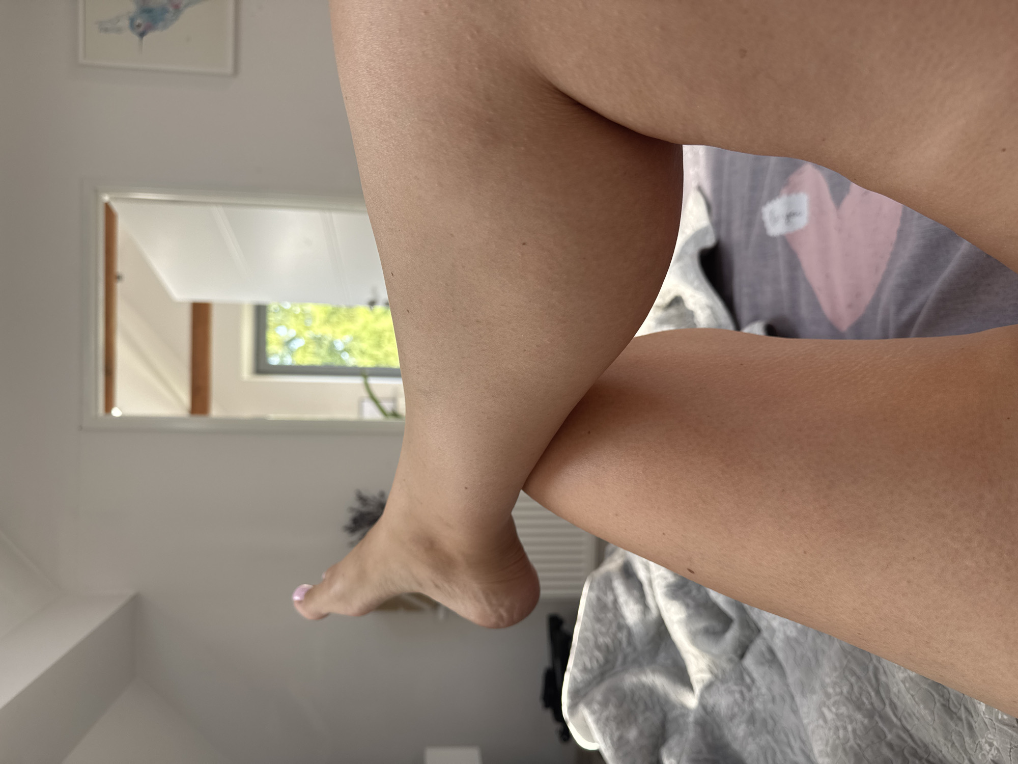 The image is a close-up, low-angle shot of a person's legs and feet, resting on a bed. The person's legs are crossed, with the left leg positioned over the right. The left foot is visible, with painted toenails, and is lifted slightly. The skin on the legs appears to have small bumps, possibly goosebumps or keratosis pilaris.

The background of the image shows a bedroom environment. The bed is covered with a grey, textured duvet or blanket. In the middle ground, there's a glimpse of a doorway leading to another room, with an open white door and a window showing greenery outside. The overall lighting suggests it's daytime, with sunlight filtering in. On the wall in the background, a framed picture of a bird is visible.

There is no information in the image to suggest this scene is located in Katowice, Poland.