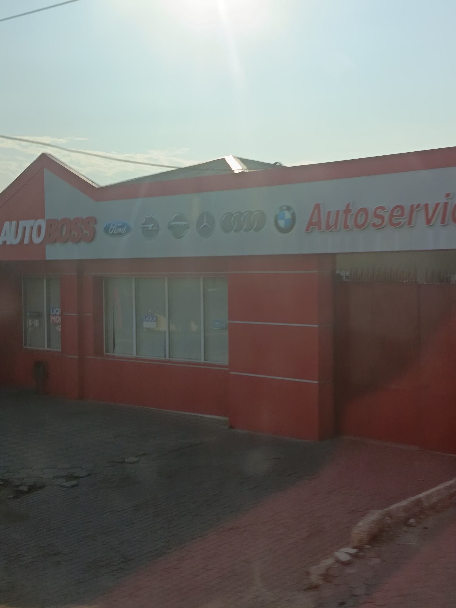 The image shows a red and white building with a sign that reads "AUTOBOSS Autoservice". Several car manufacturer logos, including Ford, Opel, Audi, and BMW, are visible on the sign. The building has several windows and a large red door on the right side. The ground in front of the building is paved with cobblestones and appears somewhat worn. The sky is bright and sunny, with some clouds visible. The scene appears to be a car service center located in Cahul, Moldova, based on information from the original image context. There are no people or vehicles clearly visible in the image.