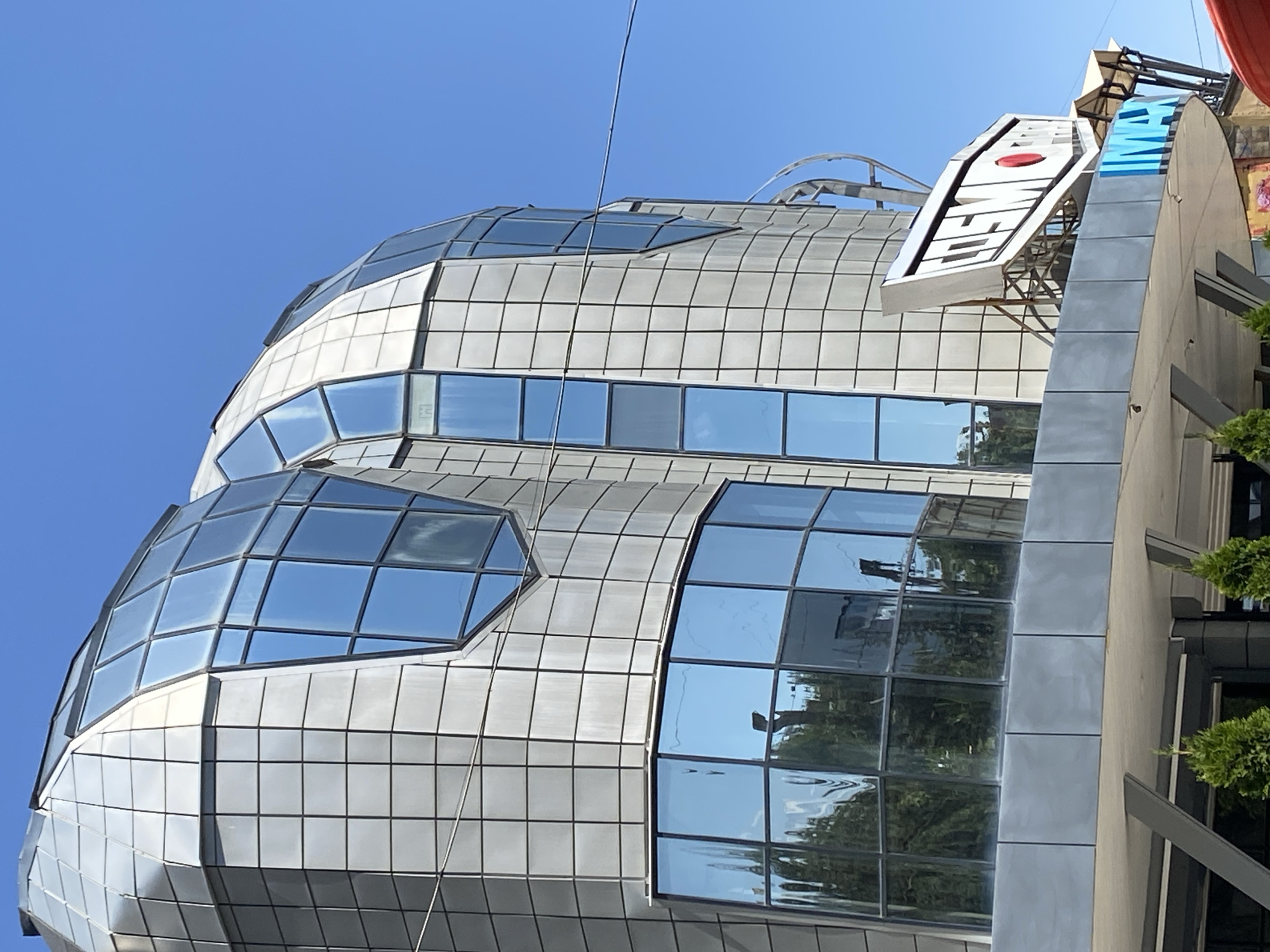 The image shows the "Cosmoport" IMAX cinema in Bishkek, Kyrgyzstan, on a clear day. The building has a distinctive modern architectural design with a curved facade covered in silver panels and large, blue-tinted glass windows. A prominent white sign with red lettering, reading "COSMOPORT" and "IMAX," is visible on the side of the building. The exterior is clean and well-maintained, and the sky is a vibrant blue. No people are visible in the frame.