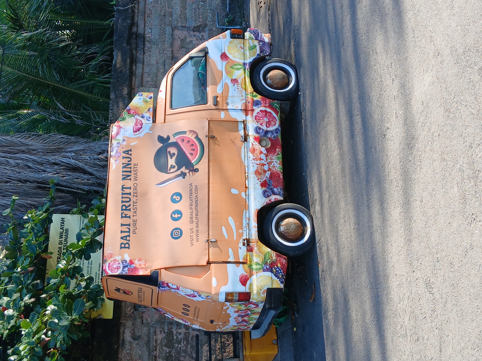 A side view of an orange food truck named "BALI FRUIT NINJA" is parked on an asphalt road in North Kuta, Indonesia. The truck is decorated with colorful graphics of various fruits, including strawberries, blueberries, oranges, lemons, and watermelons. A cartoon ninja character holding a slice of watermelon and a sword is depicted on the side of the truck, along with social media icons for Instagram, Facebook, and TikTok. The truck's text displays "PURE TASTE, ZERO WASTE" and website information. In the background, there are green trees, palm trees, and a stone wall. The ground shows shadows cast by the trees.