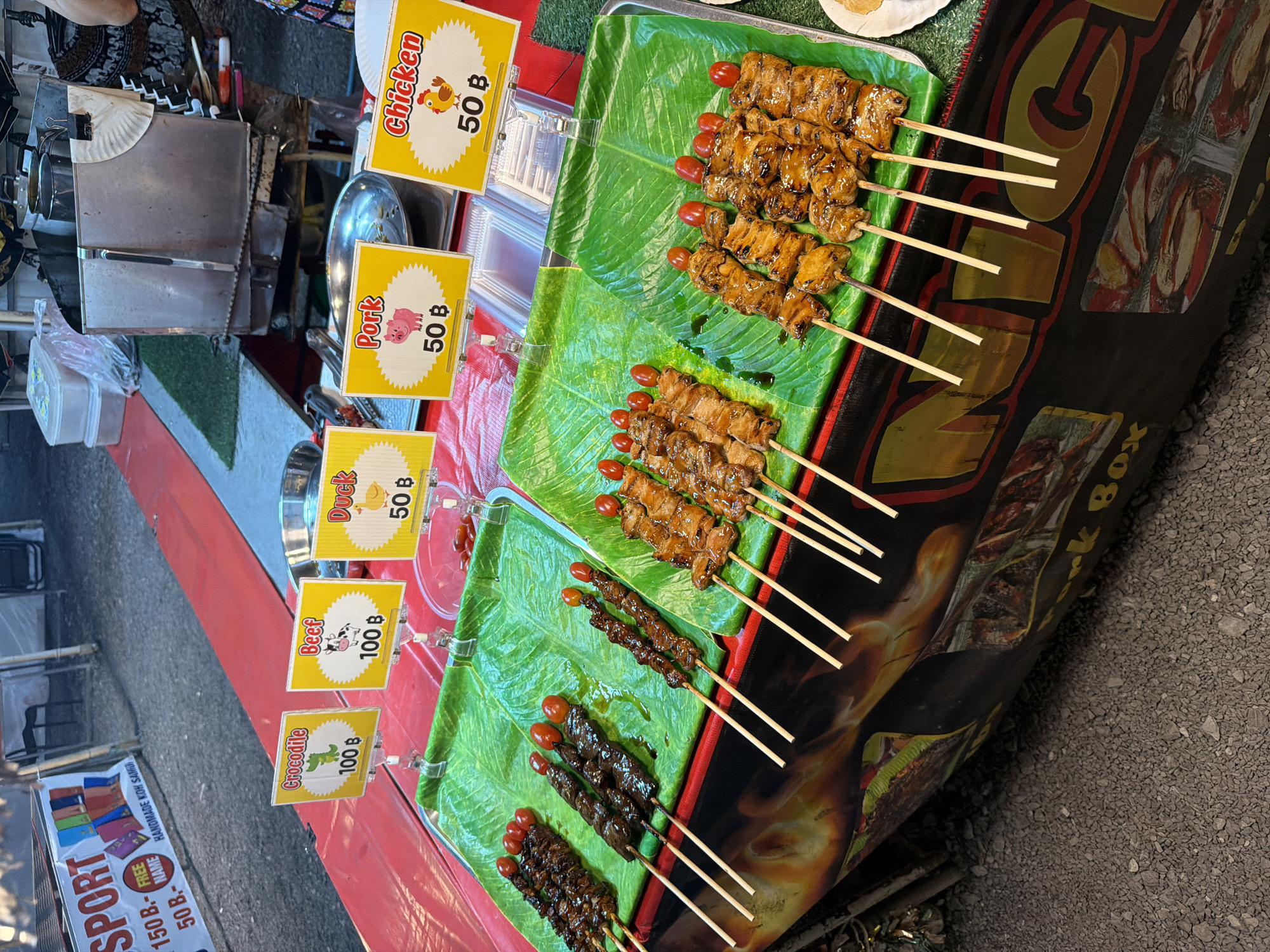 Here's a detailed description of the image:
The image shows a street food stall in Baan Bo Phut, Thailand. Various meat skewers are displayed on green trays lined with artificial banana leaves. Chicken and pork skewers cost 50 baht, while crocodile and beef skewers cost 100 baht. Each skewer is topped with a cherry tomato. The stall has signage for each meat type and a sign advertising "Sport" items from Koh Samui. The stall itself is decorated with an image of flames and the words "Nork Box".