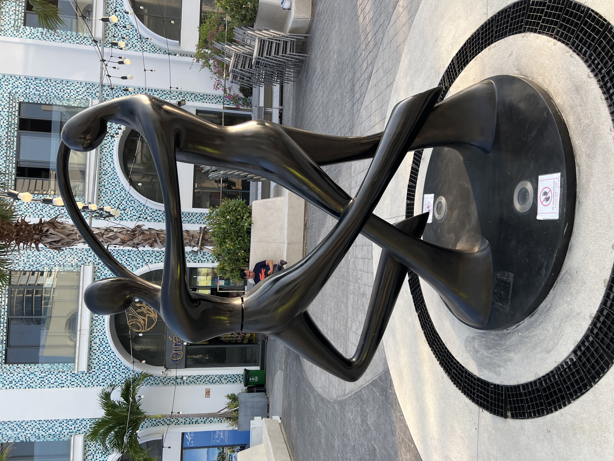 In Nha Trang, Vietnam, a contemporary sculpture of two intertwined human figures in a fluid, abstract form stands prominently in an outdoor urban plaza. The dark, polished metal sculpture, with its elongated limbs and graceful curves, evokes movement and dance. It is situated on a circular base surrounded by a grate system and a textured pavement. Behind the sculpture, a modern building with blue and white tiled facade and arched windows provides a backdrop. String lights are suspended overhead, suggesting the plaza is an area for leisure and entertainment, possibly a promenade or a public square in a resort area. A man is seated on a concrete bench to the right of the sculpture, looking at his phone. Greenery, including palm trees and bushes, adds to the tropical ambiance. Stacks of chairs are visible to the far right, indicating outdoor seating arrangements. Two small signs are affixed to the base of the sculpture, one with an icon indicating "do not step".
