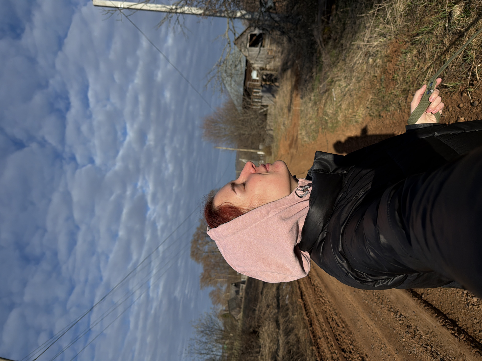 A medium shot, taken outdoors in Заболотово, Russia, shows a person from the waist up, their face tilted skyward. The person wears a black puffy jacket and a pink hooded sweatshirt. Their red hair is visible at the edge of the hood. They hold a green leash in their right hand. The background features a dirt road with tracks, dry grass, a wooden, dilapidated building, bare trees, and a sky filled with textured, layered clouds. Overhead wires cut across the upper part of the sky.