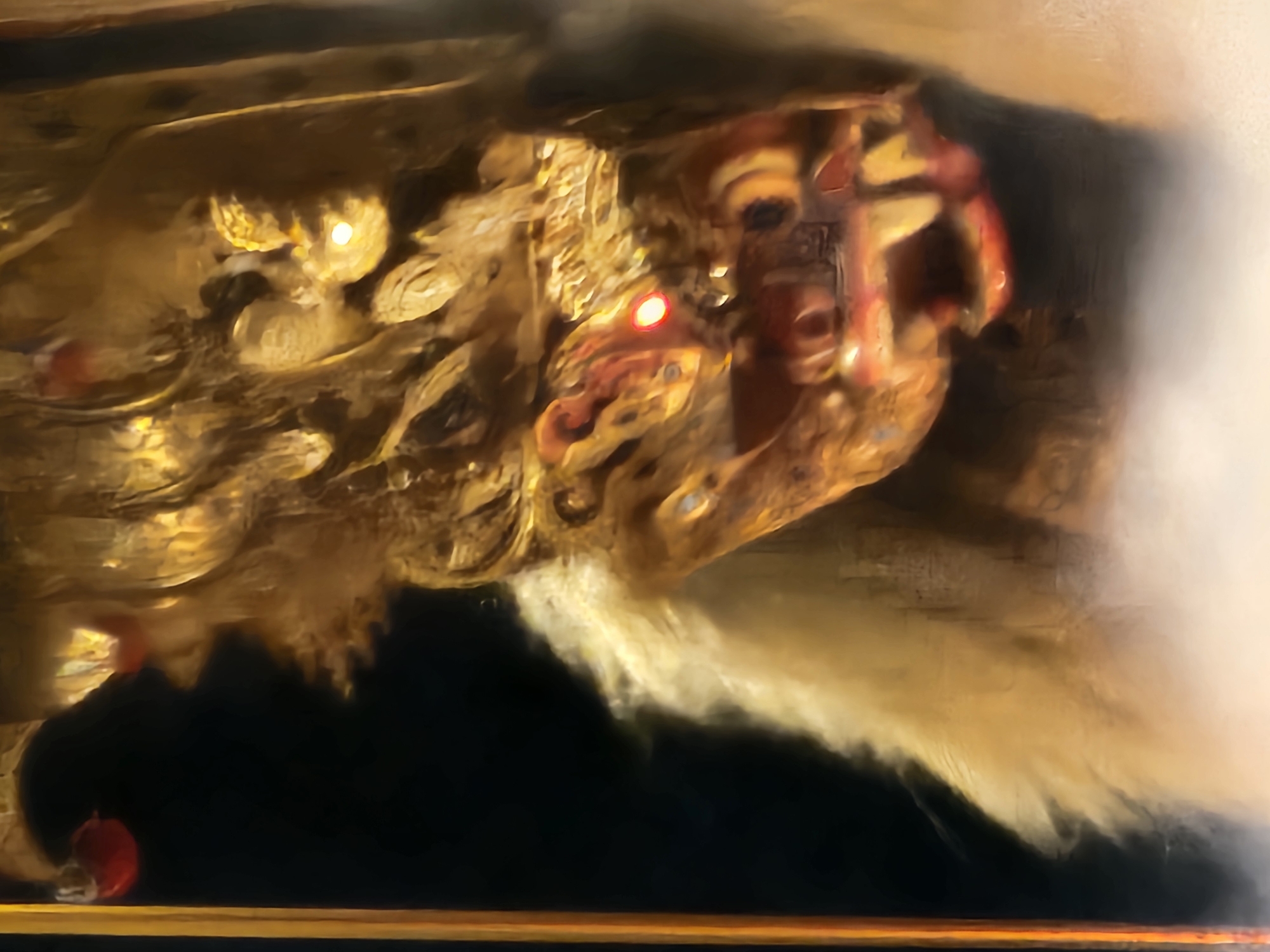The image depicts a close-up of a gilded lion-like figure, possibly part of a larger structure or tableau. The figure has intricate detailing and appears to be illuminated by lights, with some appearing red. The surrounding environment is dark, with the lower portion of the image showing what appears to be a horizontal surface. The composition and style of the image are suggestive of a cultural or religious artifact in Indonesia.