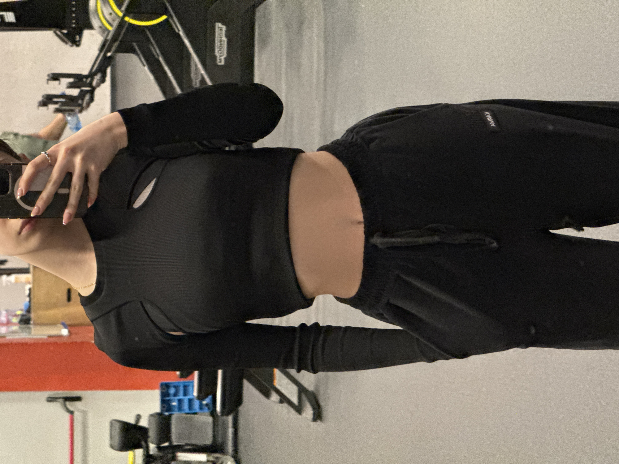 A person wearing a black long-sleeved crop top and black sweatpants is taking a selfie in what appears to be a gym. Gym equipment is visible in the background. The exact location in Sofia, Bulgaria is not discernible from the image.