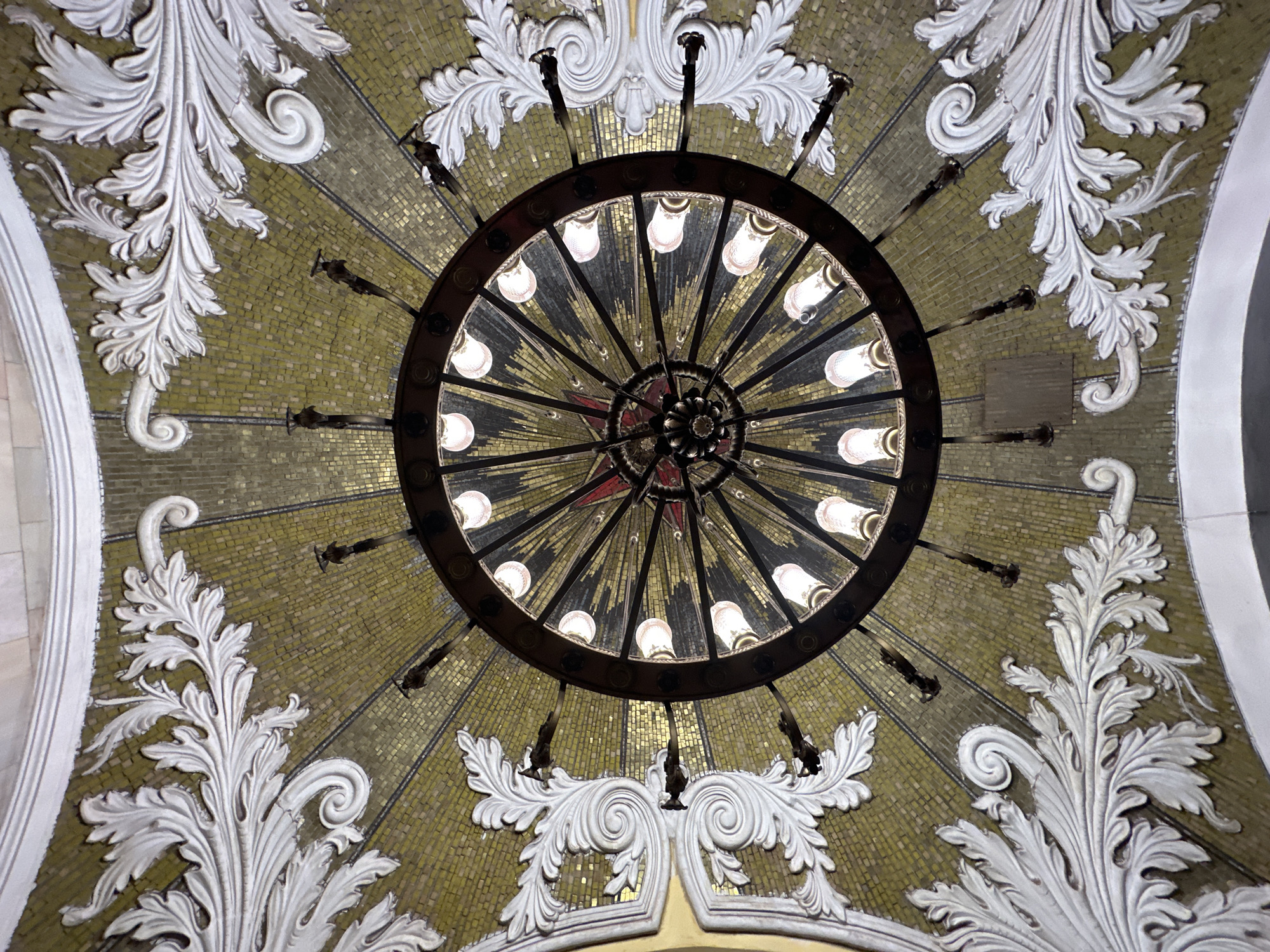 The image depicts an ornate ceiling inside the Komsomolskaya-Koltsevaya metro station in Moscow, Russia. A large, circular chandelier with multiple light bulbs hangs in the center, adorned with a red star at its core. The ceiling is covered in golden-brown mosaic tiles, arranged in a radial pattern emanating from the center. Decorative white plaster floral motifs are distributed across the ceiling's surface, framing the chandelier and mosaic background. The lighting is a combination of the light bulbs from the chandelier and the ambient light reflecting off the ceiling.